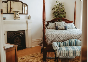A bedroom features a four-poster bed with patterned beeeing and pillows, a decorative fireplace with a mantelpiece clock and mirror above it, and a striped chaise lounge in front of the bed.