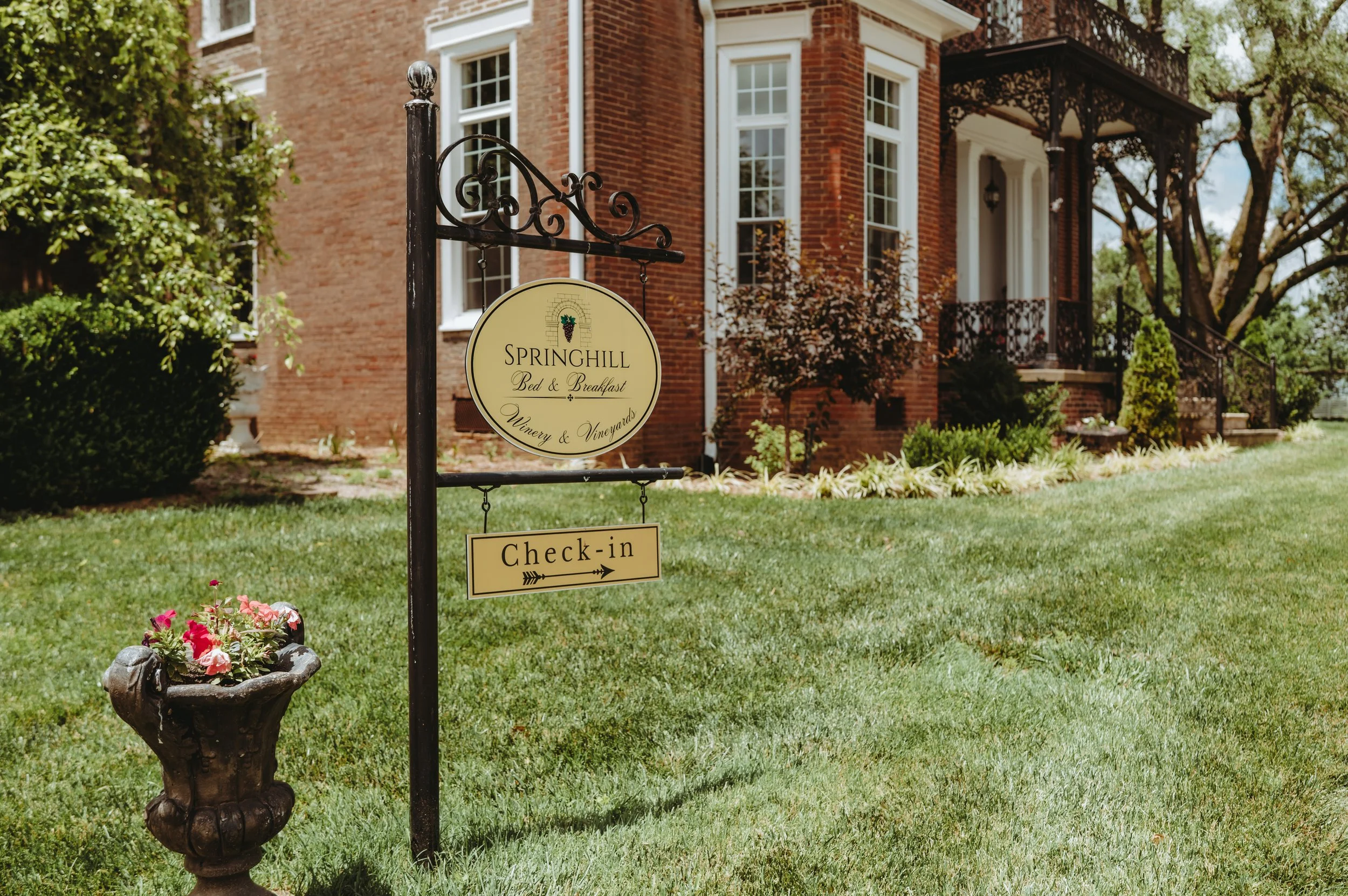 Brick house with white-trim windows and a covered porch, surrounded by green trees and a manicured lawn; a decorative signpost stands in the foreground near flower beds.