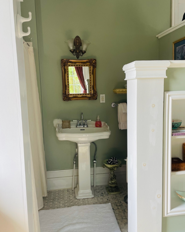 A pedestal sink with a gold framed mirror above it in a bathroom with pale green walls and a tiled floor.