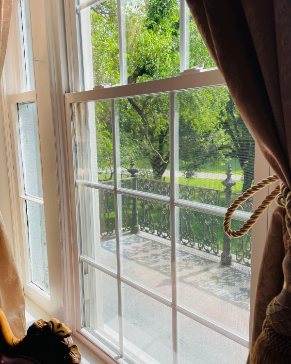 Elegant Rose Suite window framed by tassel curtains, overlooking a wrought‑iron railing and sunlit trees.