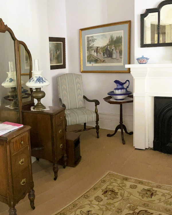 Sitting area with striped upright chair and table with basin and pitcher; two framed pictures hang above on a gray wall. White fireplace with mirror above. Vanity dresser with lamp in foreground. Cream rug on the floor.