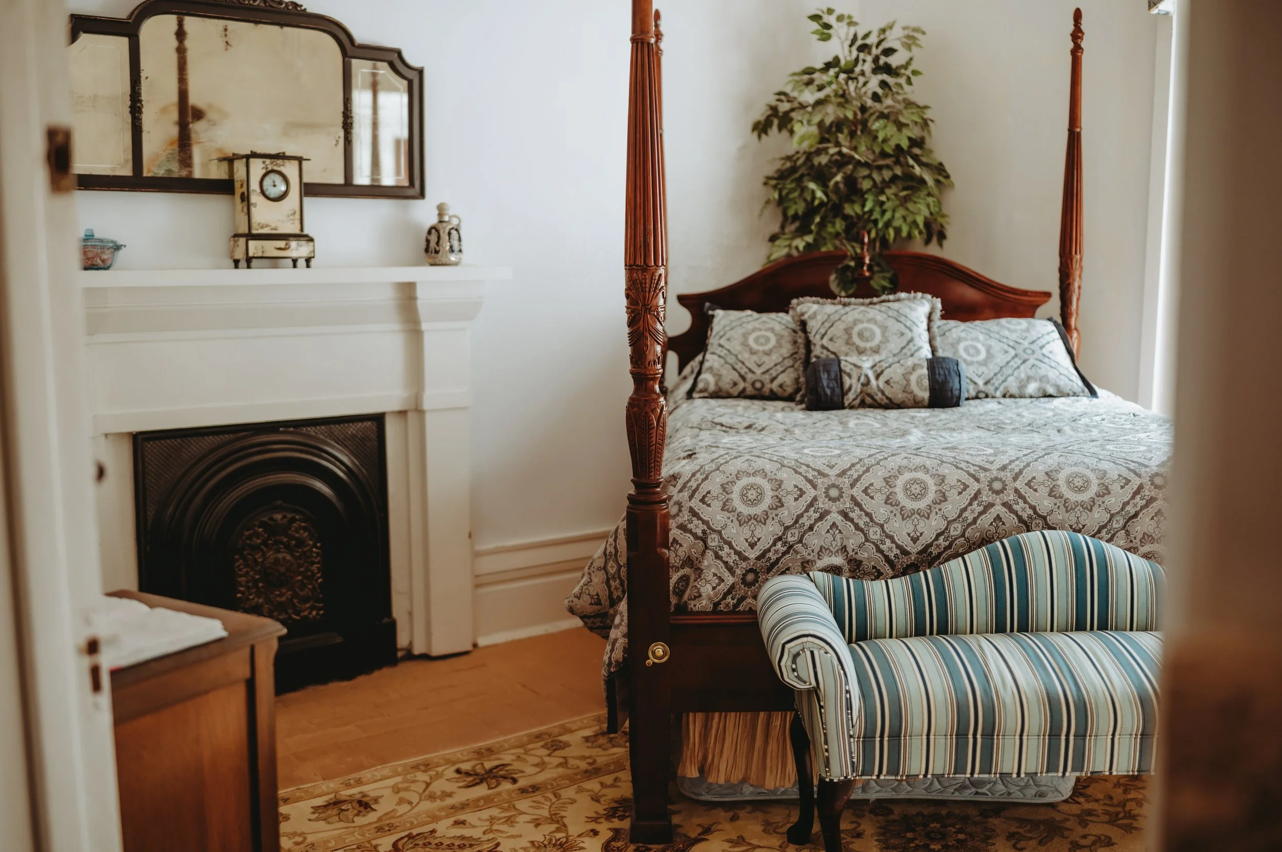 A bedroom features a four-poster bed with patterned bedding and pillows, a decorative fireplace with a mantelpiece clock and mirror above it, and a striped chaise lounge i front of the bed