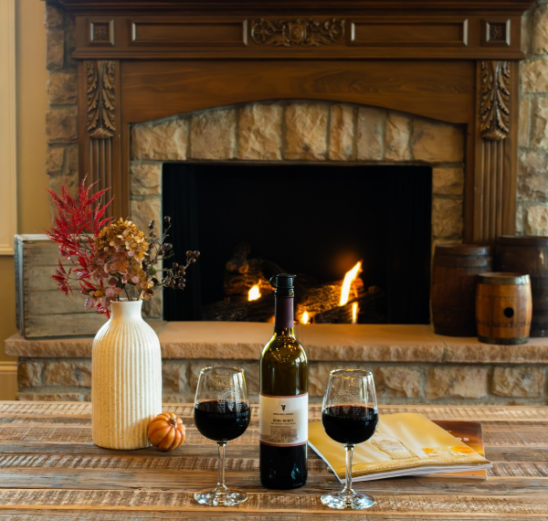 A coffee table in front of a stone fireplace with two filled wine glasses, a bottle, a vase of red and pink flowers, and warm firelight in the hearth.