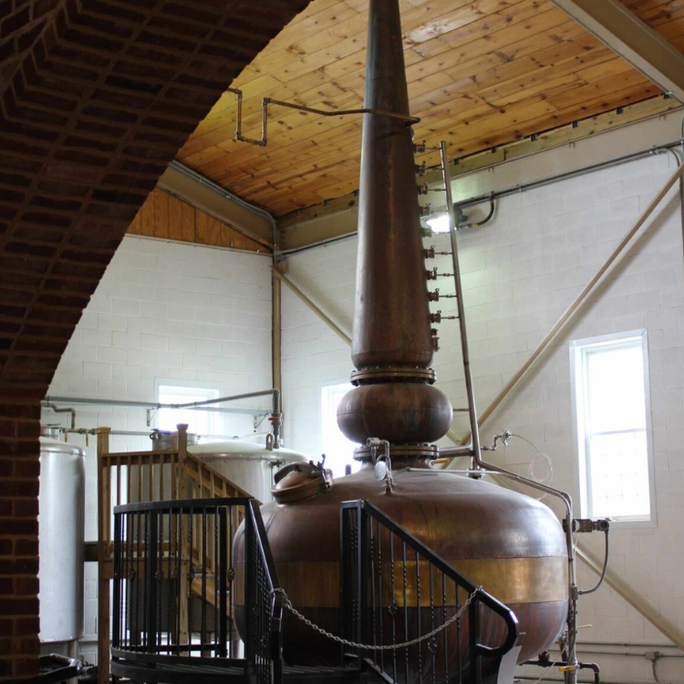 Large copper still inside a distillery, with a wooden ceiling and white brick walls, surrounded by pipes and equipment.