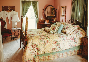 A bedroom featuring a four-poster bed with floral comforter, a red armchair, a dresser with an oval mirror, and two white robes hanging on a rack.
