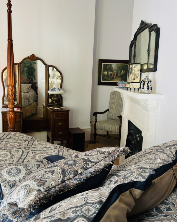 A four-poster bed in foreground with patterned comforter and pillows. Vanity table with mirror sets against a gray wall; striped upright chair to the right with framed picture above. White fireplace mantel with objects set on top and mirror above.