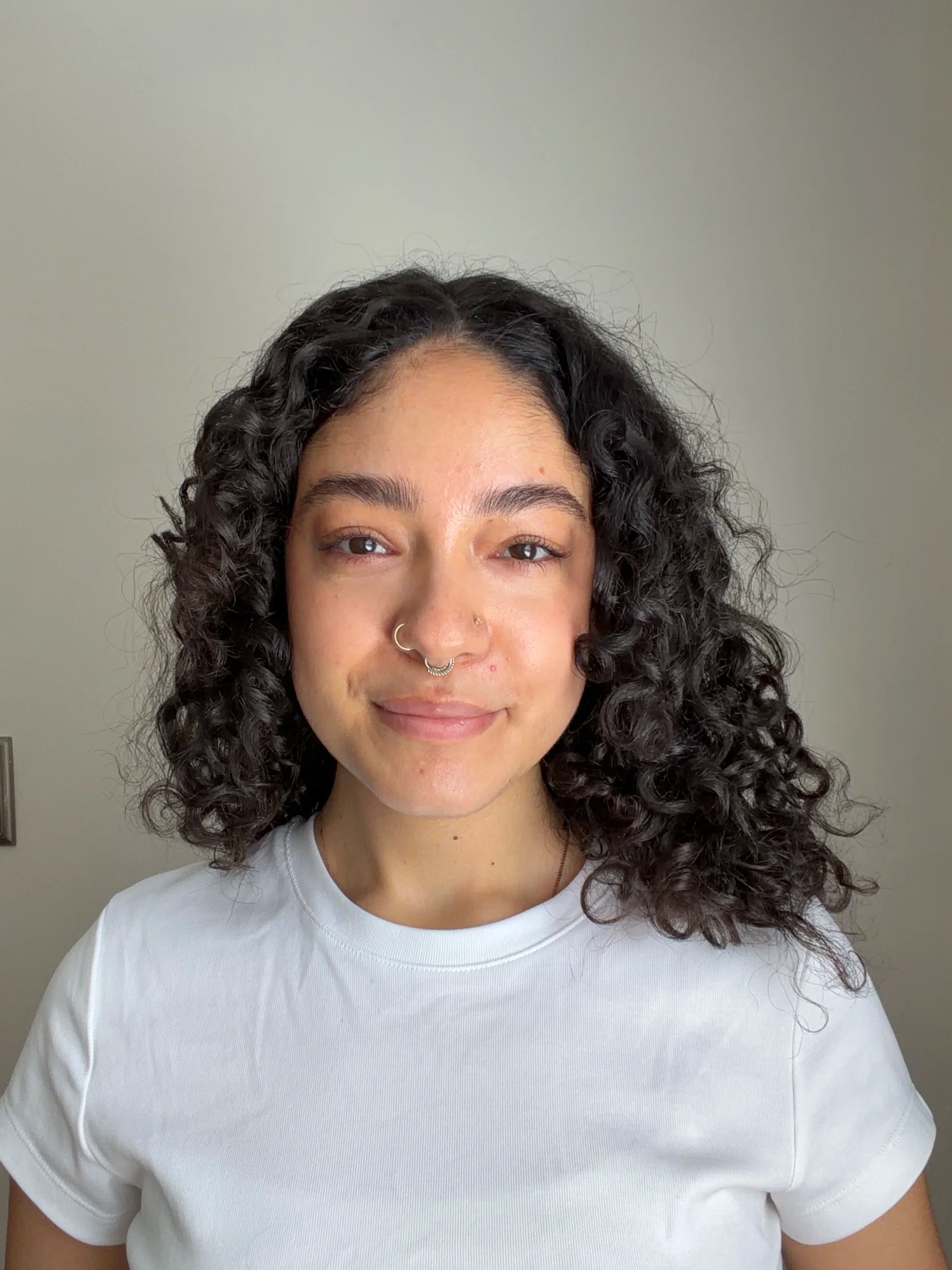 Close-up of a woman with curly dark hair wearing a white t-shirt, smiling against a plain light-colored background.
