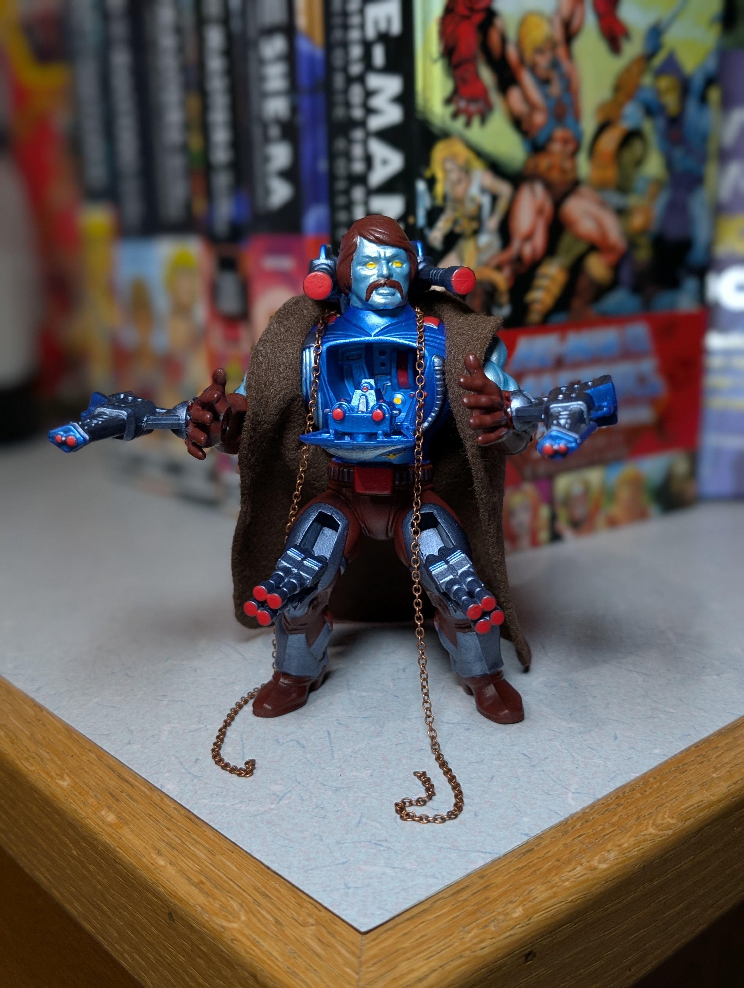 Action figure of a blue-skinned character with brown hair and beard dressed in a sci-fi outfit, wearing a brown cape, with weapons in hands, standing on a wooden surface with comic books in the background.