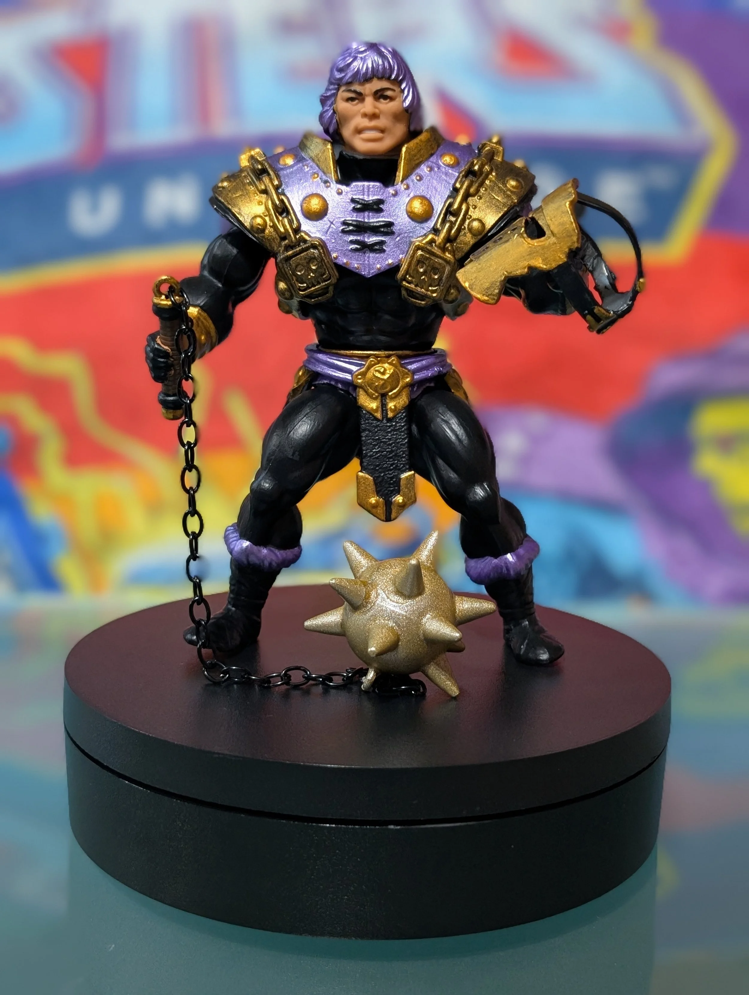 Action figure of a muscular warrior with purple hair, wearing black and gold armor, holding a chain in one hand and standing on a spiked gold ball, displayed on a black circular stand.