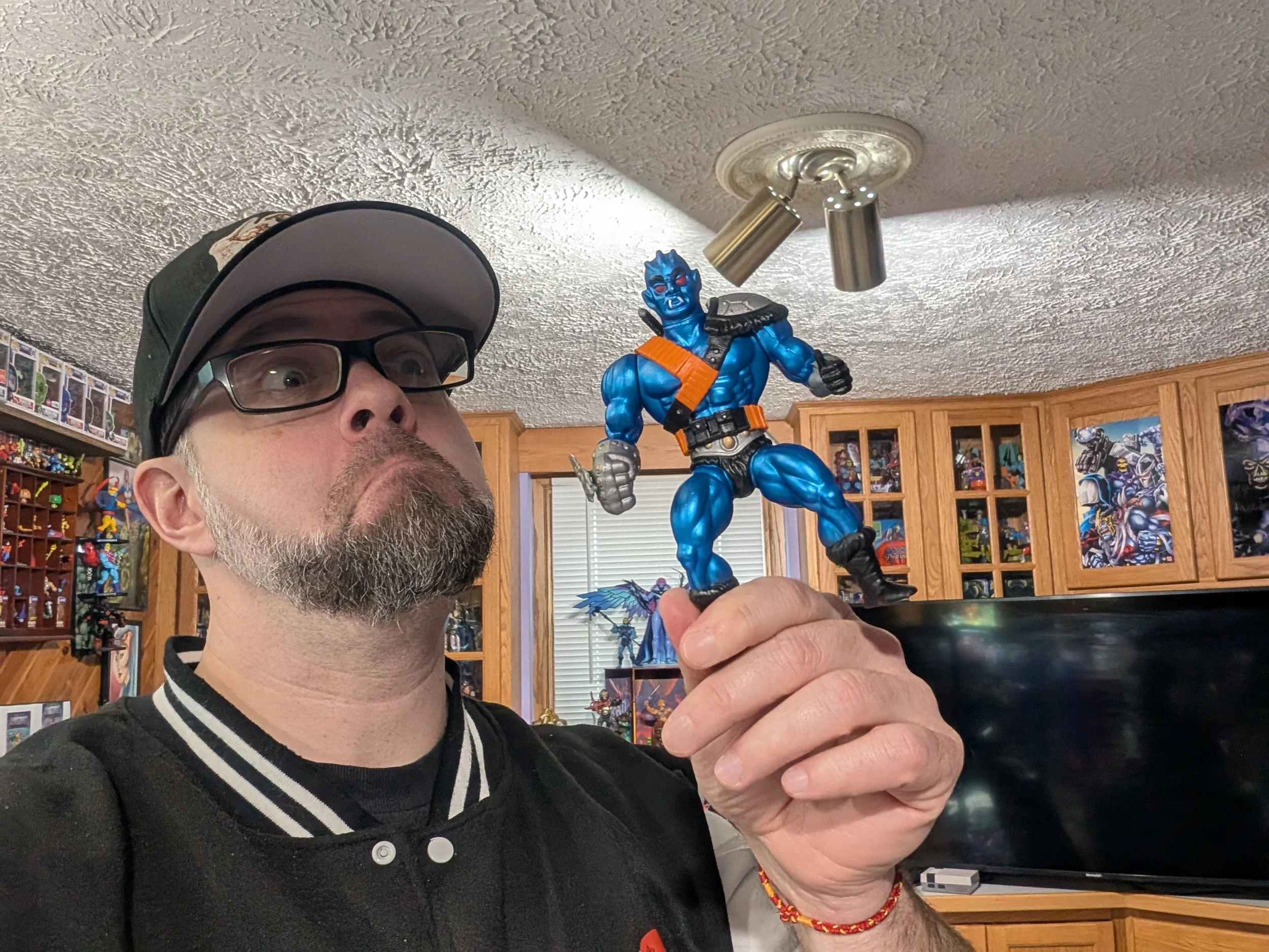 A man with glasses and a black and white jacket holding a blue action figure of a muscular, blue-skinned character with red eyes, a mischievous expression, and a black and orange outfit. The background shows a room with wooden cabinets, shelves with collectibles, and framed artwork.
