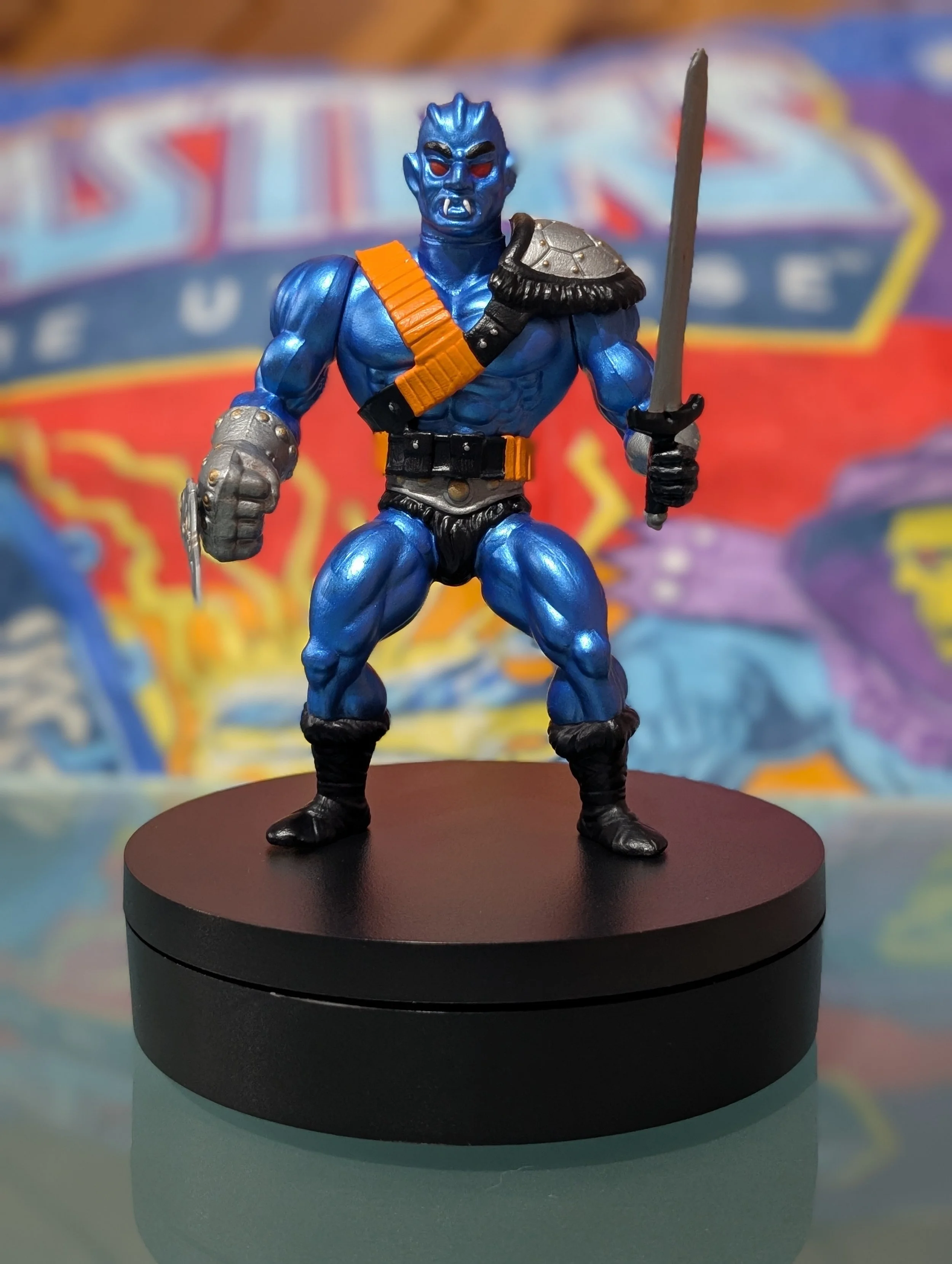 A blue-skinned action figure with red eyes, fangs, and pointed ears, dressed in a black loincloth, with orange and gray armor accents, holding a sword in one hand and a shield on its shoulder, standing on a round black display stand.