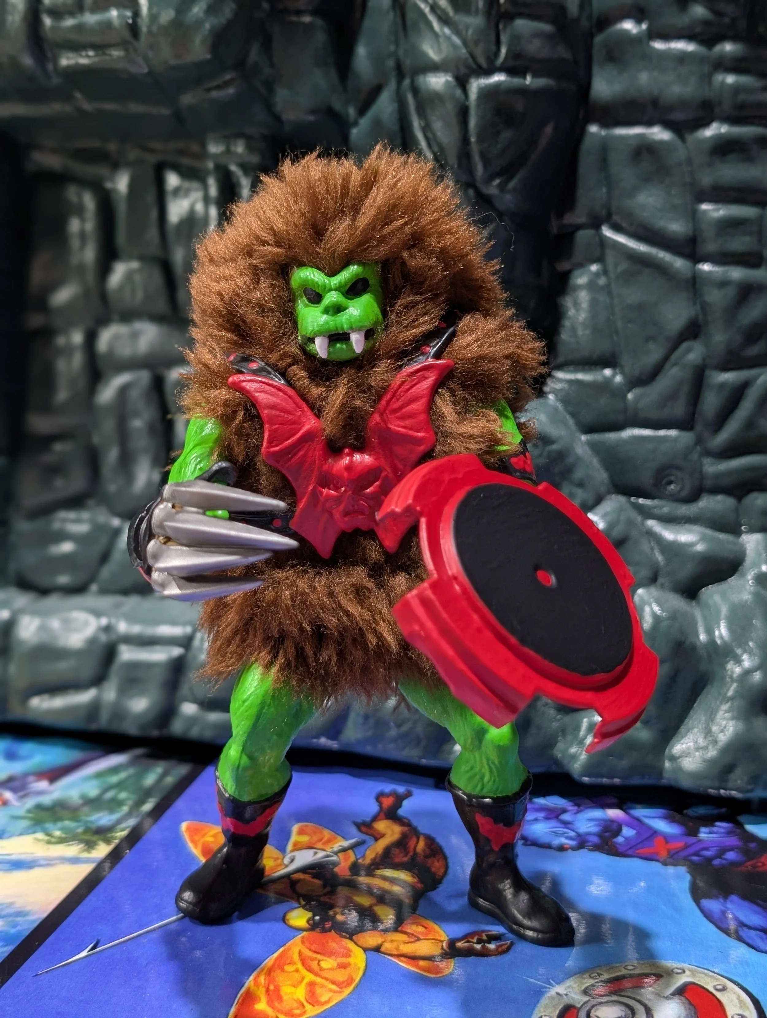 Action figure of a green creature with a fierce face, brown hair, red armor, and black boots, holding a weapon. The background showcases a dark, rocky setting.
