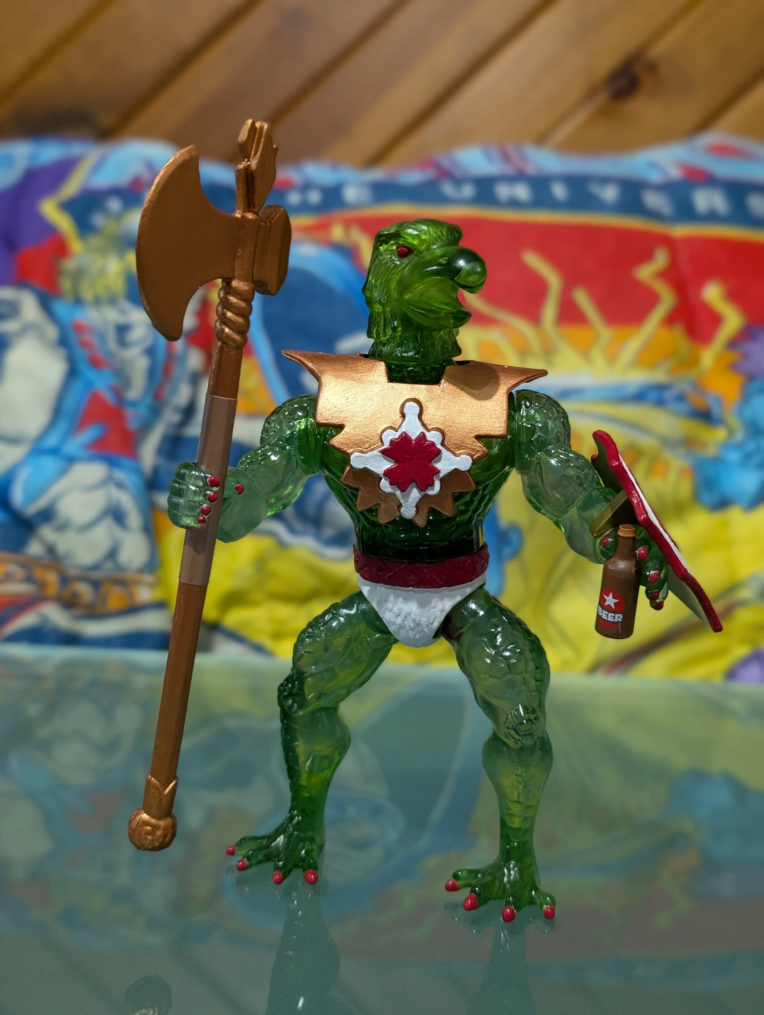 A toy figure of a green monster with red eyes, wearing a gold star-shaped medal, holding an axe and a bottle of beer, standing on a reflective surface with a colorful cloth background.