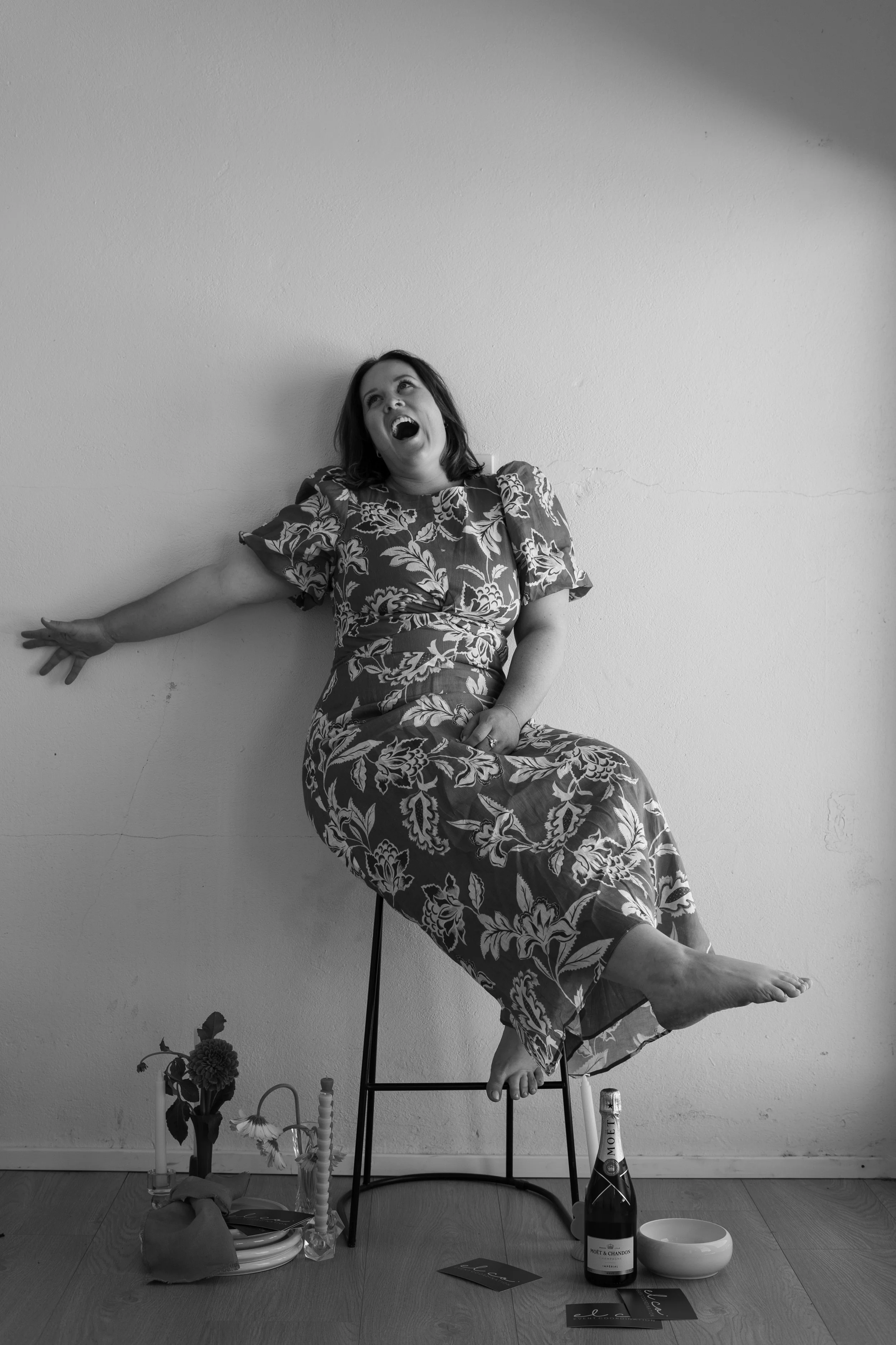 A woman with dark hair wearing a floral dress is sitting on a stool against a plain wall, appearing to laugh or sing loudly with her mouth wide open. Objects including a flower in a vase, a bottle of Moët & Chandon champagne, a bowl, magazines, and a few cards are arranged on the floor in front of her.