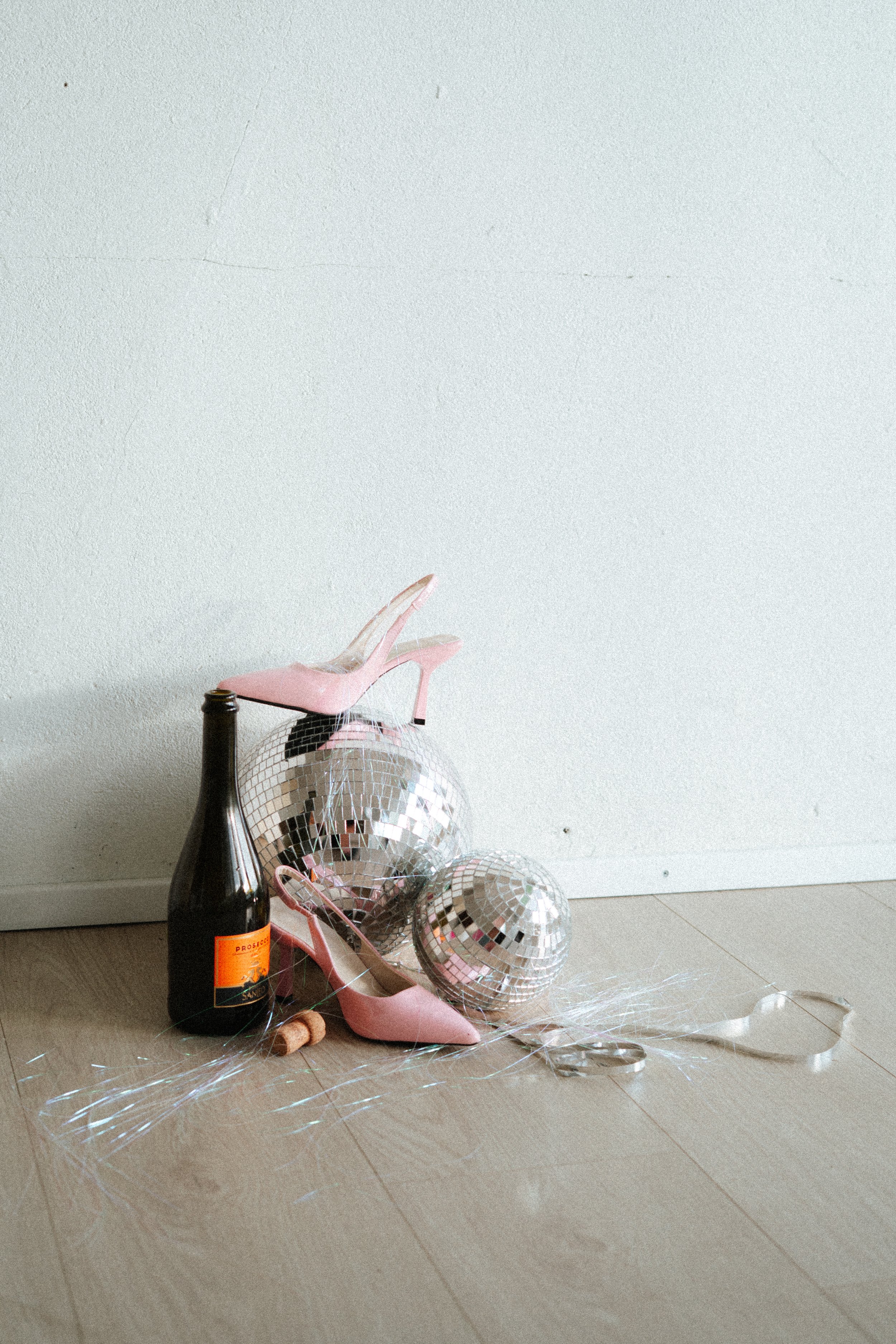 Pink high-heeled shoe, disco balls, a bottle of wine with a bright orange label, small cork, and silver tinsel on a beige tiled floor against a plain white wall.