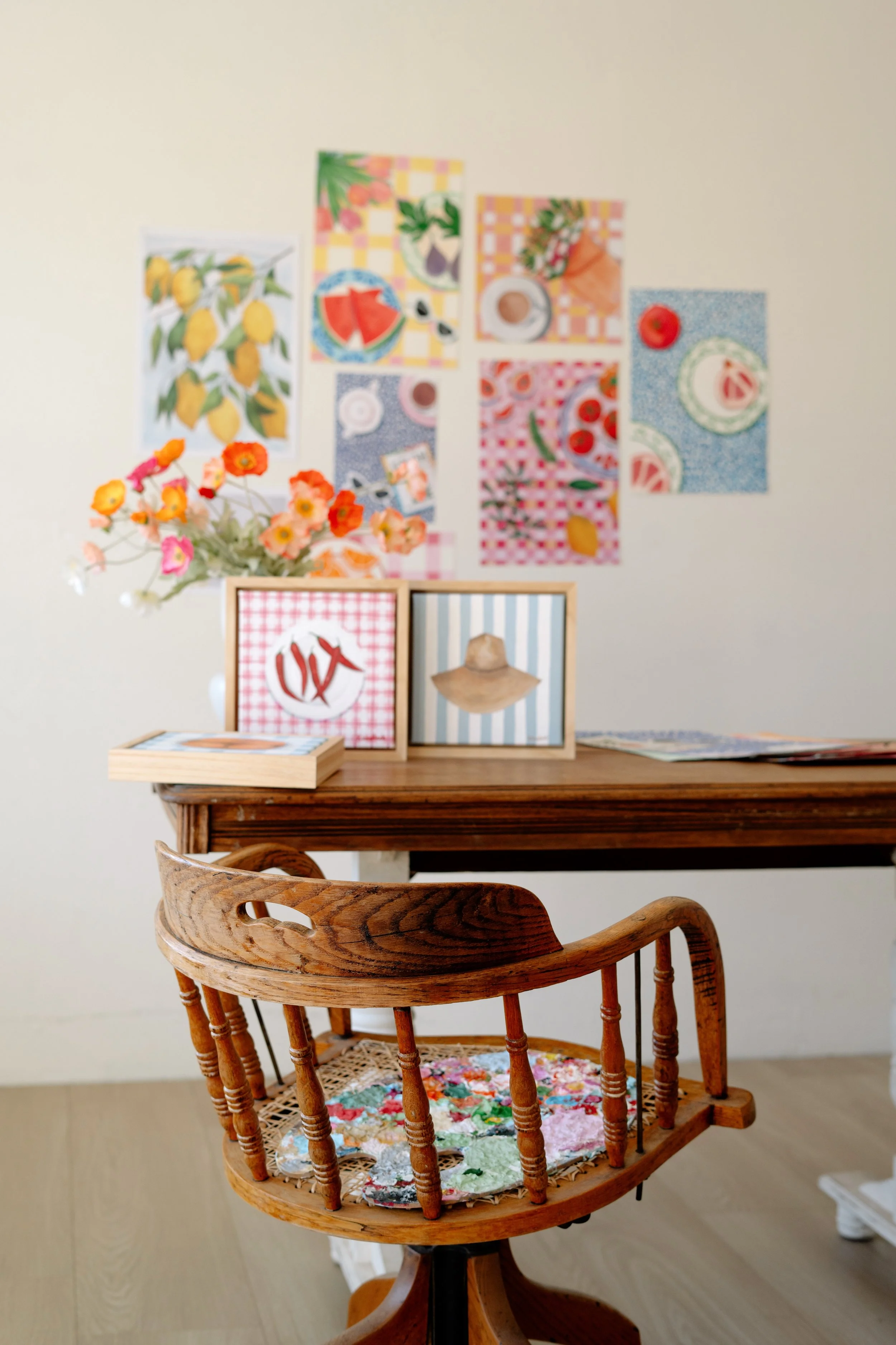 A wooden table with framed artwork and a vase of pink and orange flowers, positioned in front of a wall decorated with colorful fruit-themed posters.
