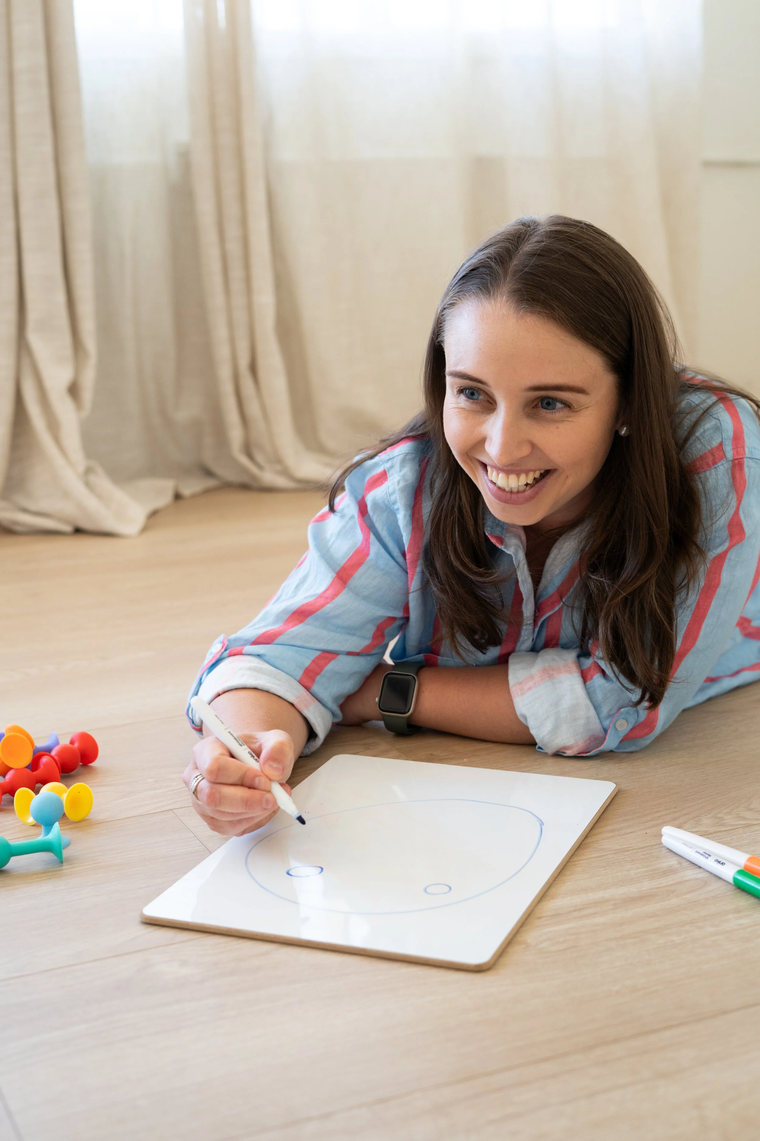 A woman with brown hair and blue eyes, wearing earrings, a plaid shirt, and a smartwatch, lies on the floor smiling, drawing on a whiteboard with colorful game pieces scattered nearby, in a room with beige curtains.