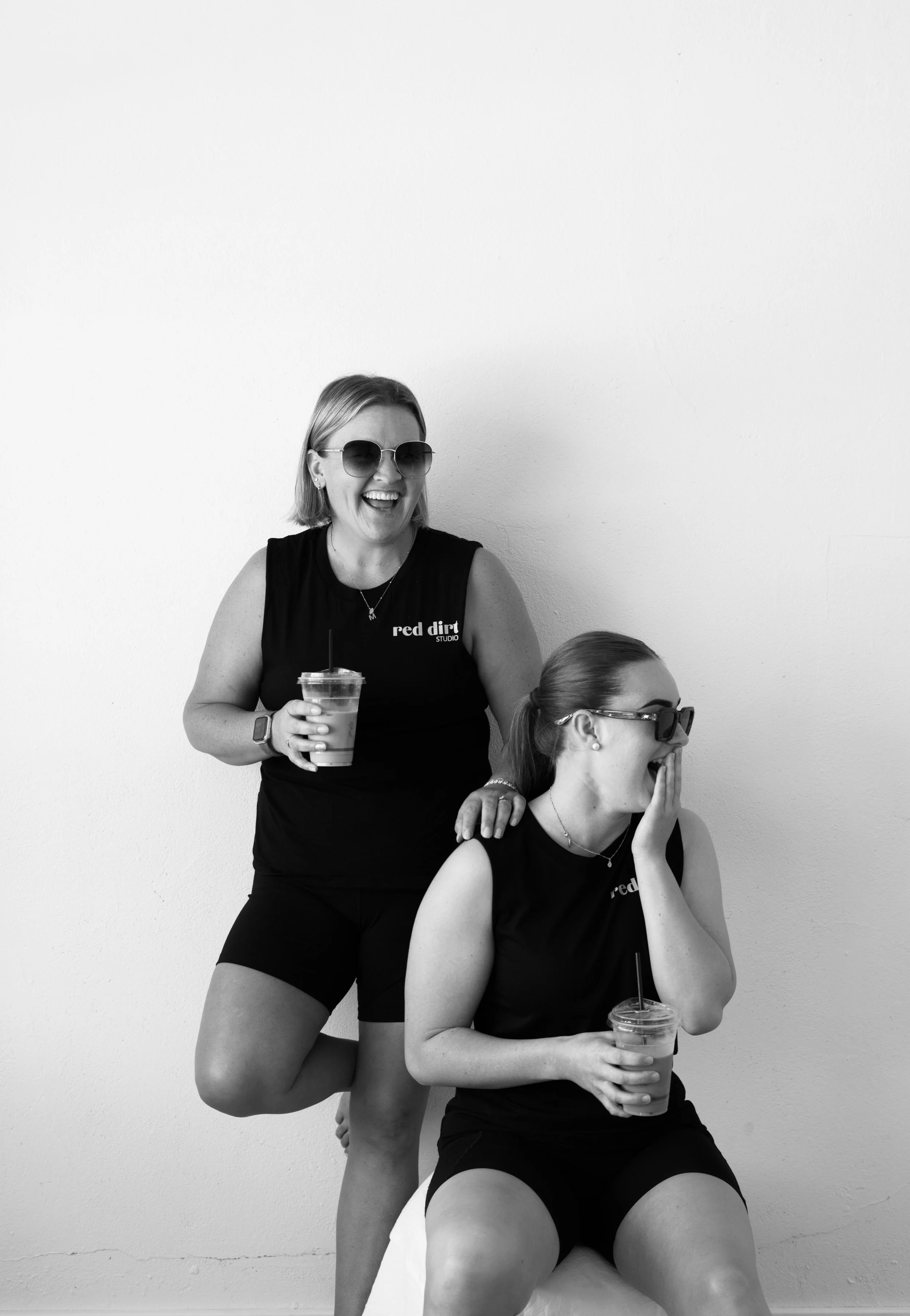 Three women in black tank tops and sunglasses enjoying drinks and laughter against a plain white wall. One woman is seated, holding a drink, with her hand over her face, while the others are standing or leaning nearby, smiling and engaging in a shared humorous moment.