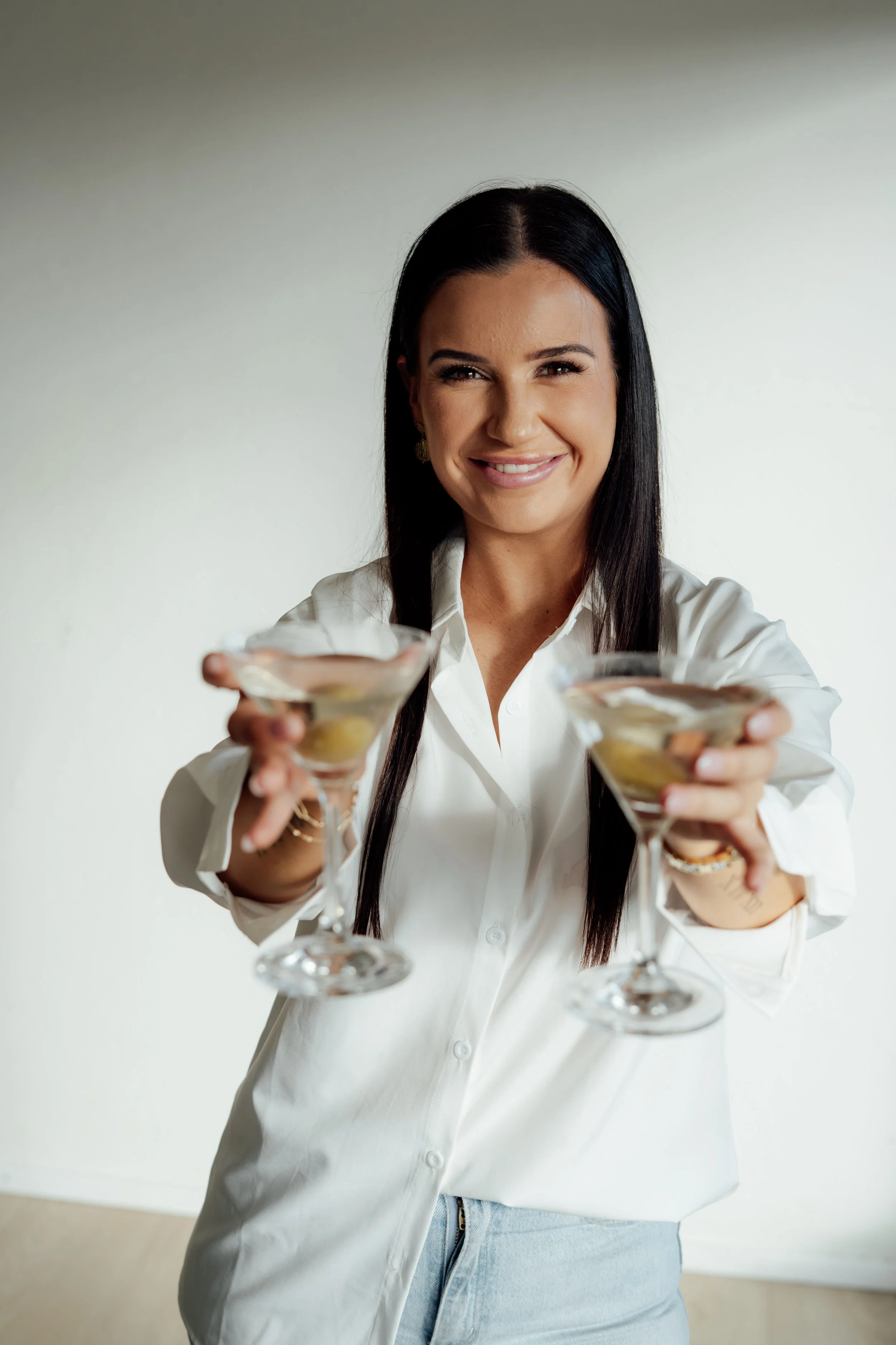 Woman with long dark hair wearing a white button-up shirt and light blue jeans, smiling and holding two martini glasses with olives, standing against a plain light-colored wall.