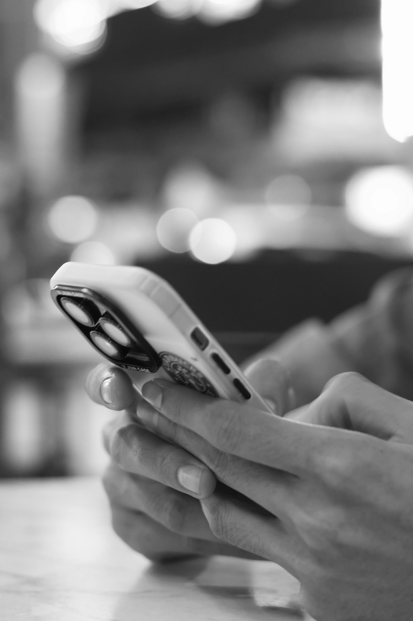 A black-and-white photograph of a person holding a smartphone with both hands, with focus on the hands and phone, and a blurred background of lights.
