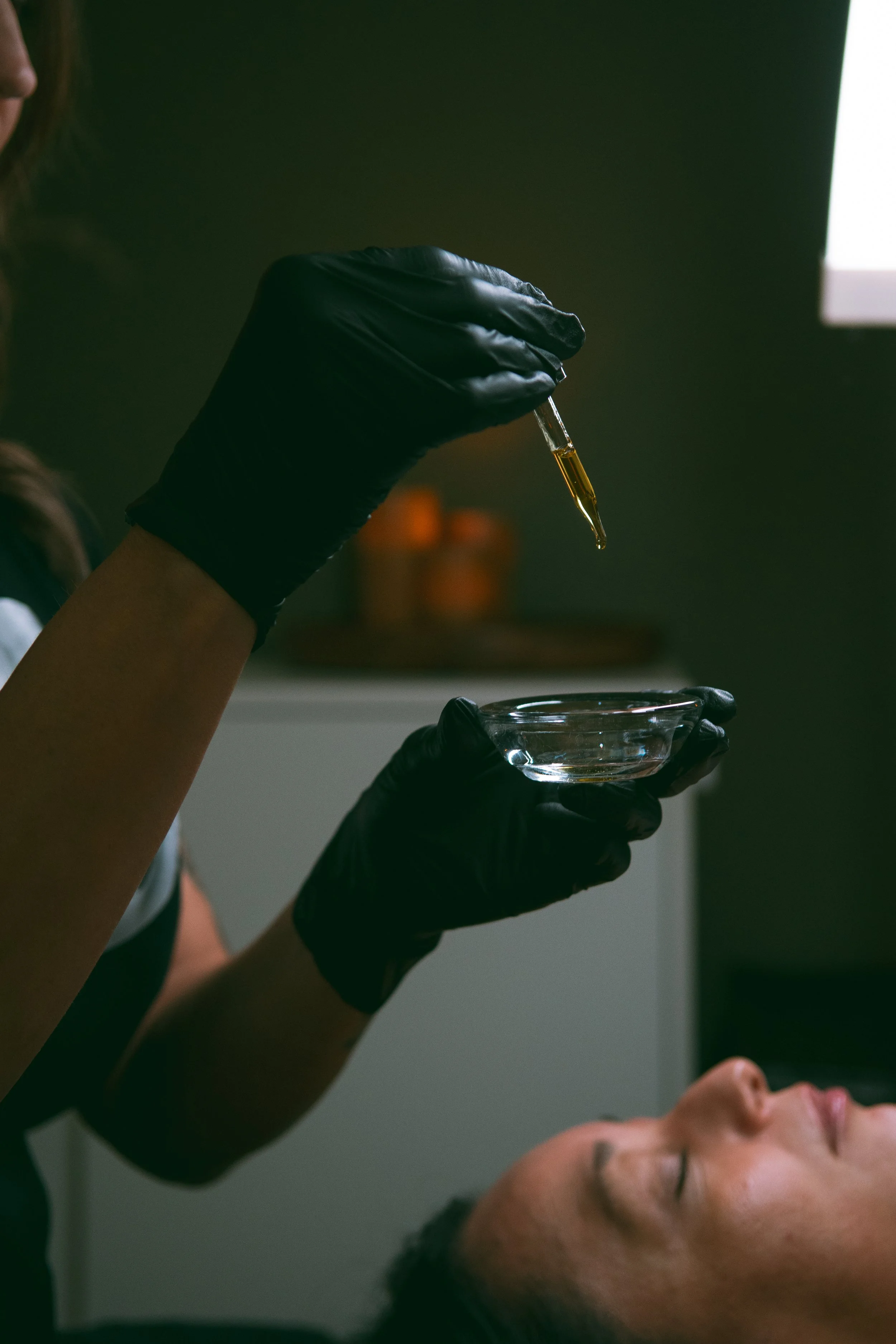 A person lying on their back with eyes closed, receiving a liquid treatment from another individual wearing black gloves. The person with gloves is using a dropper to place a liquid into a small glass bowl.