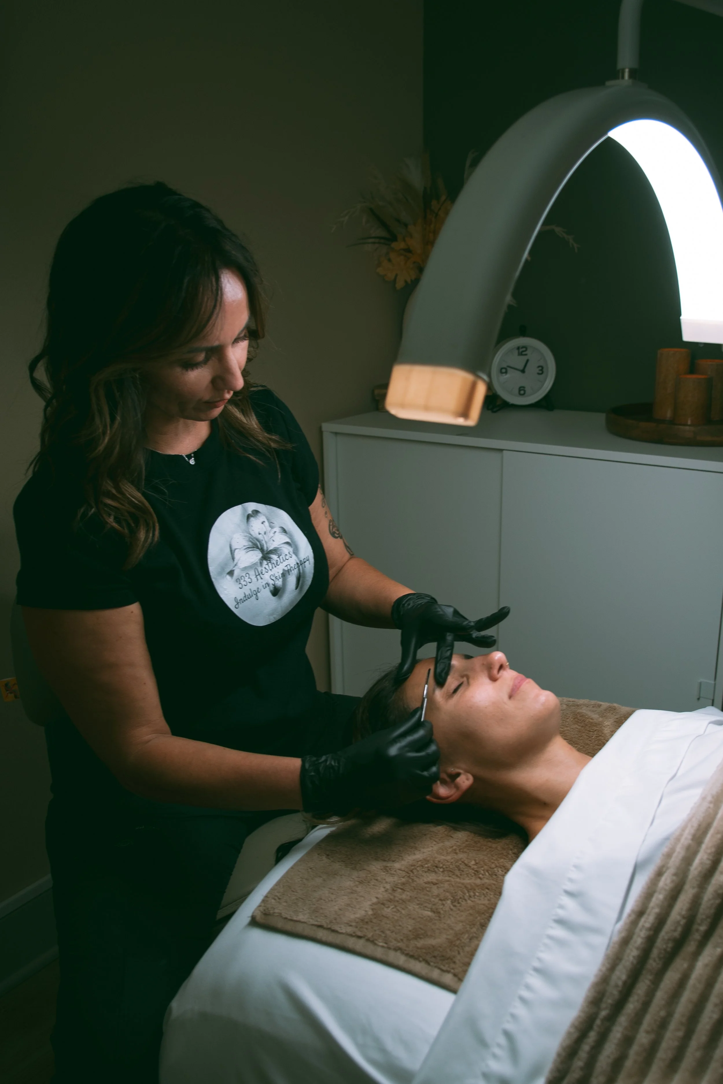 A woman receiving a facial treatment from a technician in a spa or aesthetic clinic, with the technician wearing black gloves.