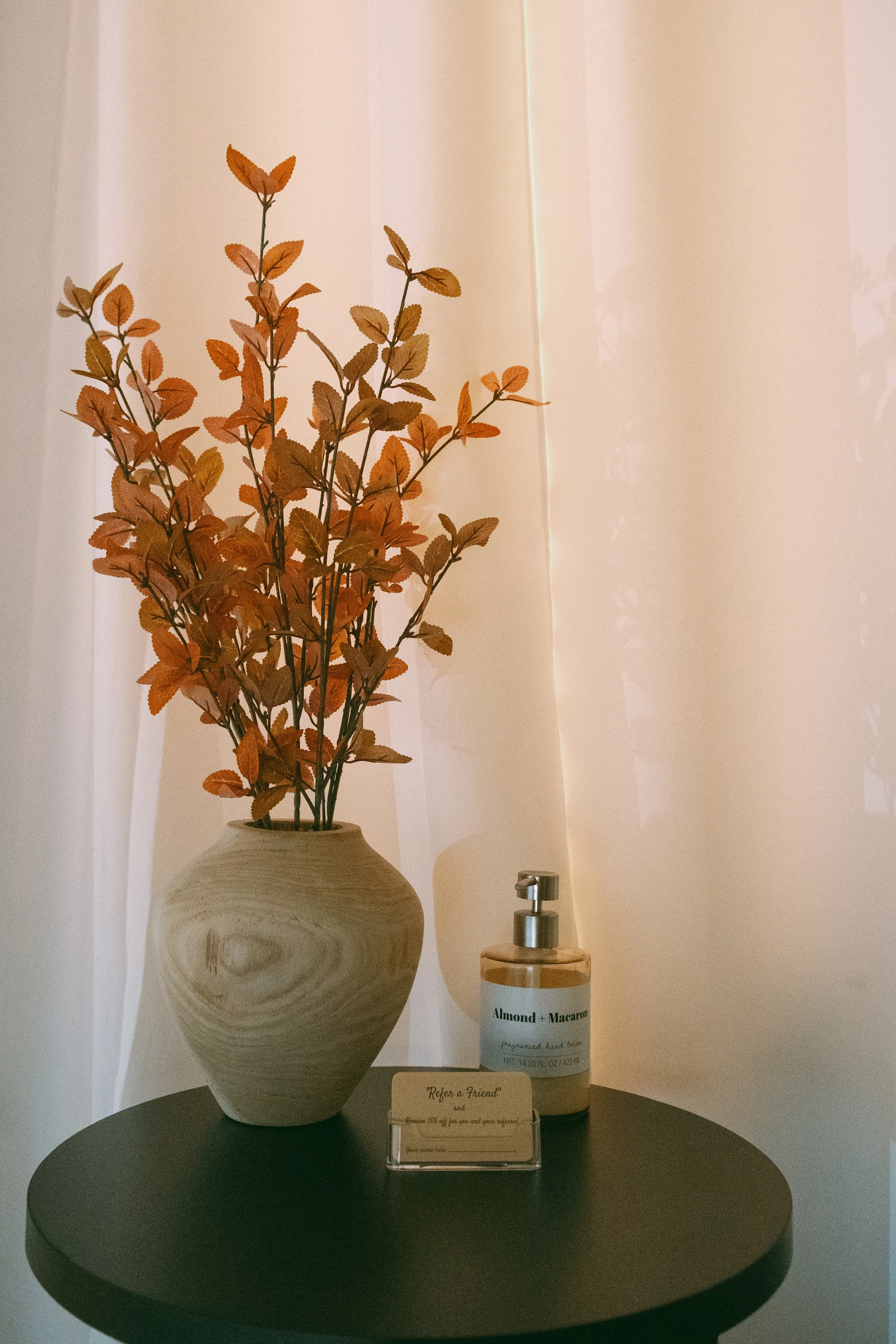 A decorative scene with a beige ceramic vase with a woodgrain pattern holding dried orange leaves, a white pump bottle labeled 'Almond + Macaron' scented hand lotion, and a small beige card with the words 'Refer a friend' on a round black table.