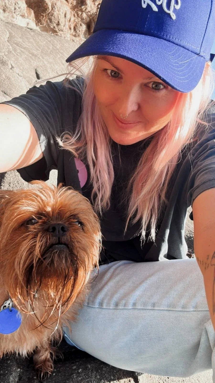 A woman with pink hair wearing a blue cap and black t-shirt taking a selfie with her dog outdoors.