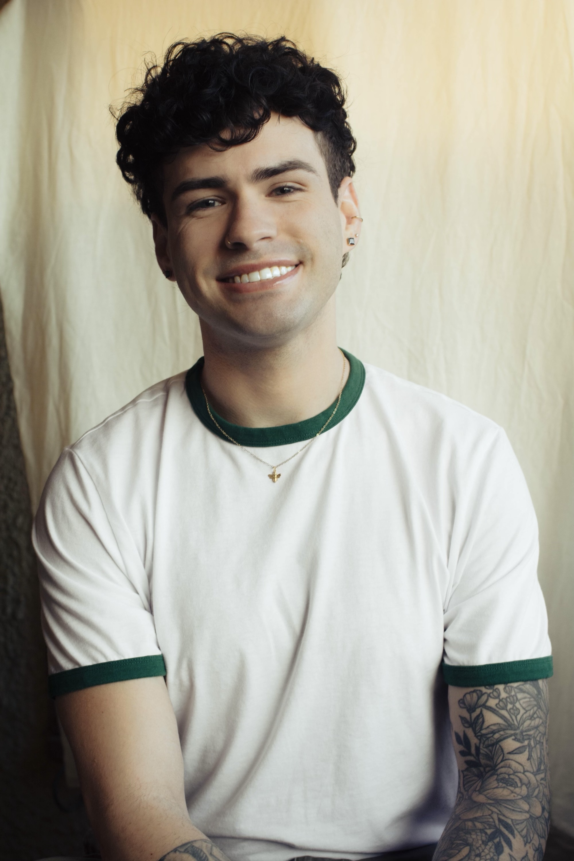 A young man with curly dark hair smiling, wearing a white t-shirt with green accents, jewelry, and tattoos on his arm, against a cream-colored curtain background.