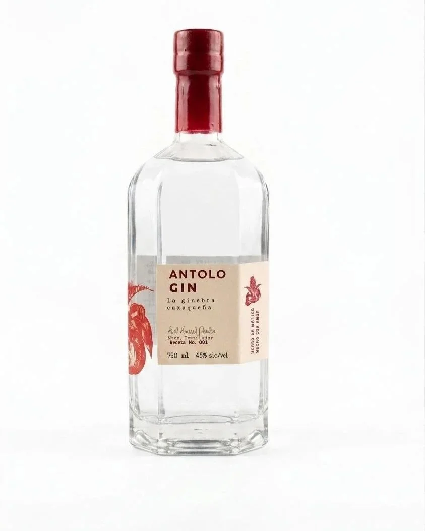 A clear glass bottle with a red cap and a beige label that reads "Antolo Gin" and "La ginebra caxaqueña" including illustrations of a red fish and a red flower.