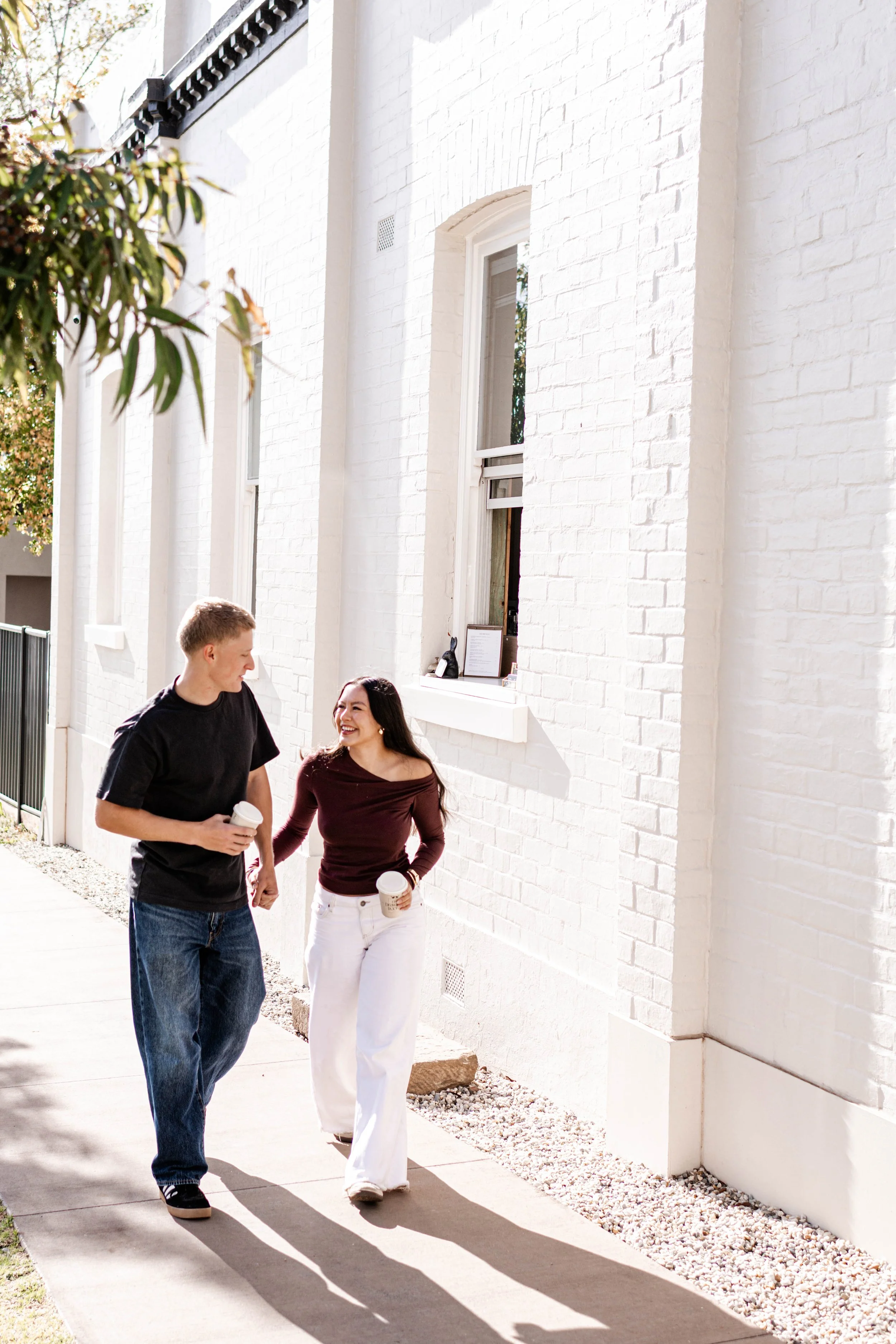 Two young adults, a man and a woman, walking and talking with coffee cups in hand outside on a sunny day, beside a white-painted brick building with large windows.