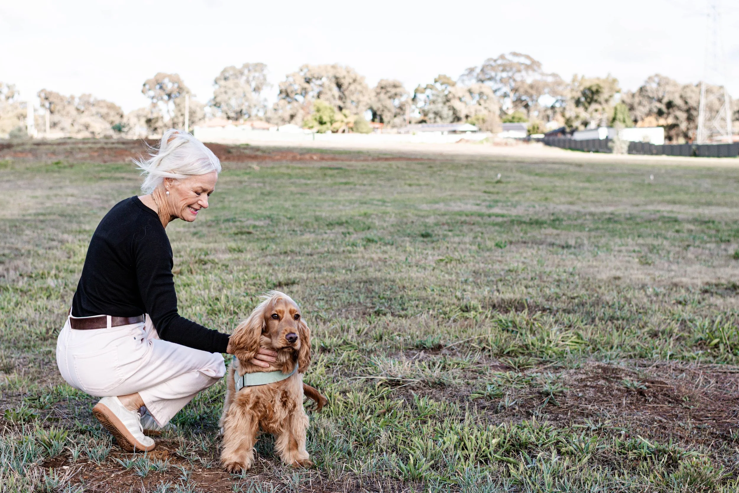 An older woman with white hair and a black top squatting in a grassy field, holding a golden retriever puppy with a harness, with trees and a fence in the background.