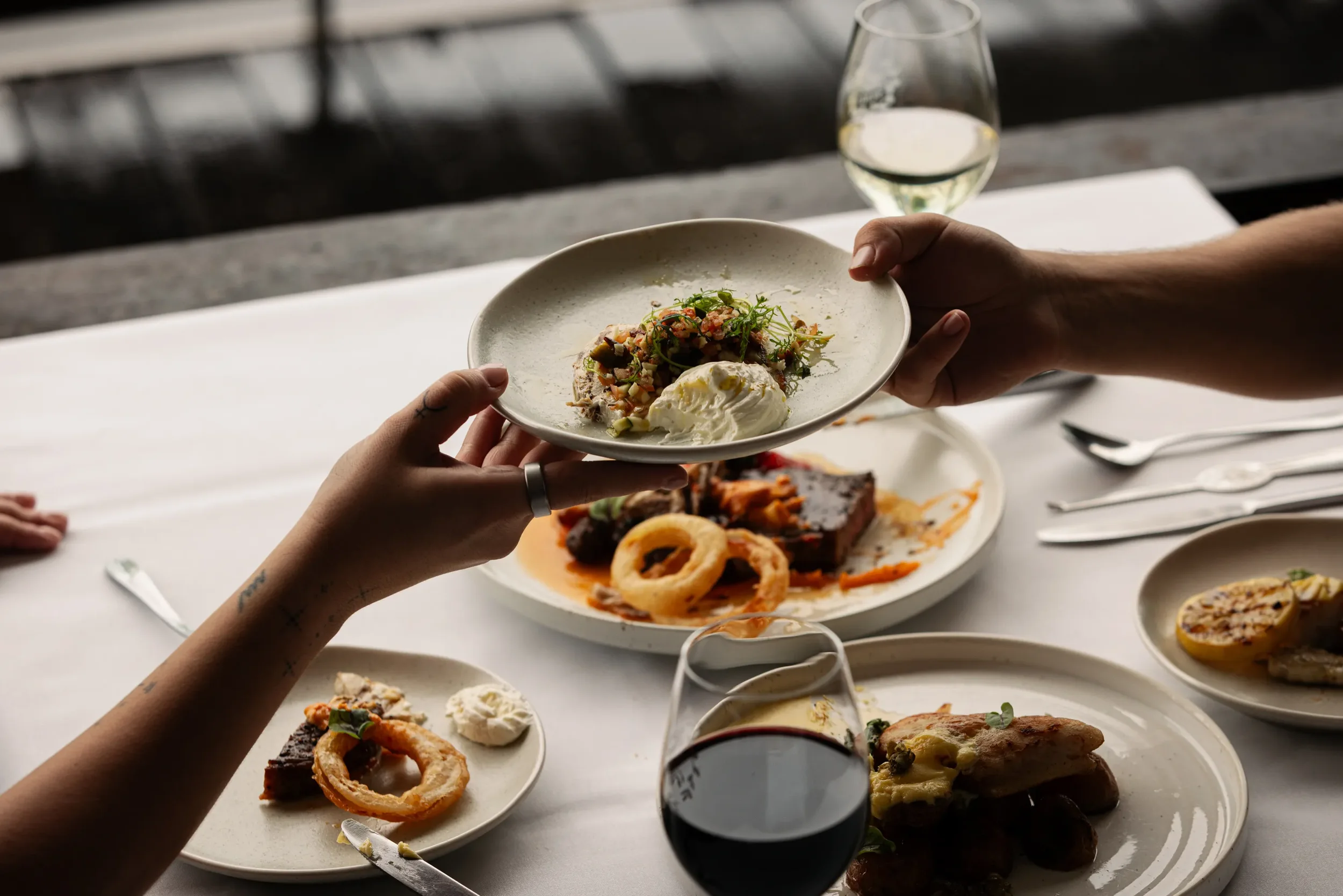 Person holding a white plate with a portion of food, including a salad, a dollop of cream, and garnishes, at a dining table with other plates of food and glasses of red and white wine.