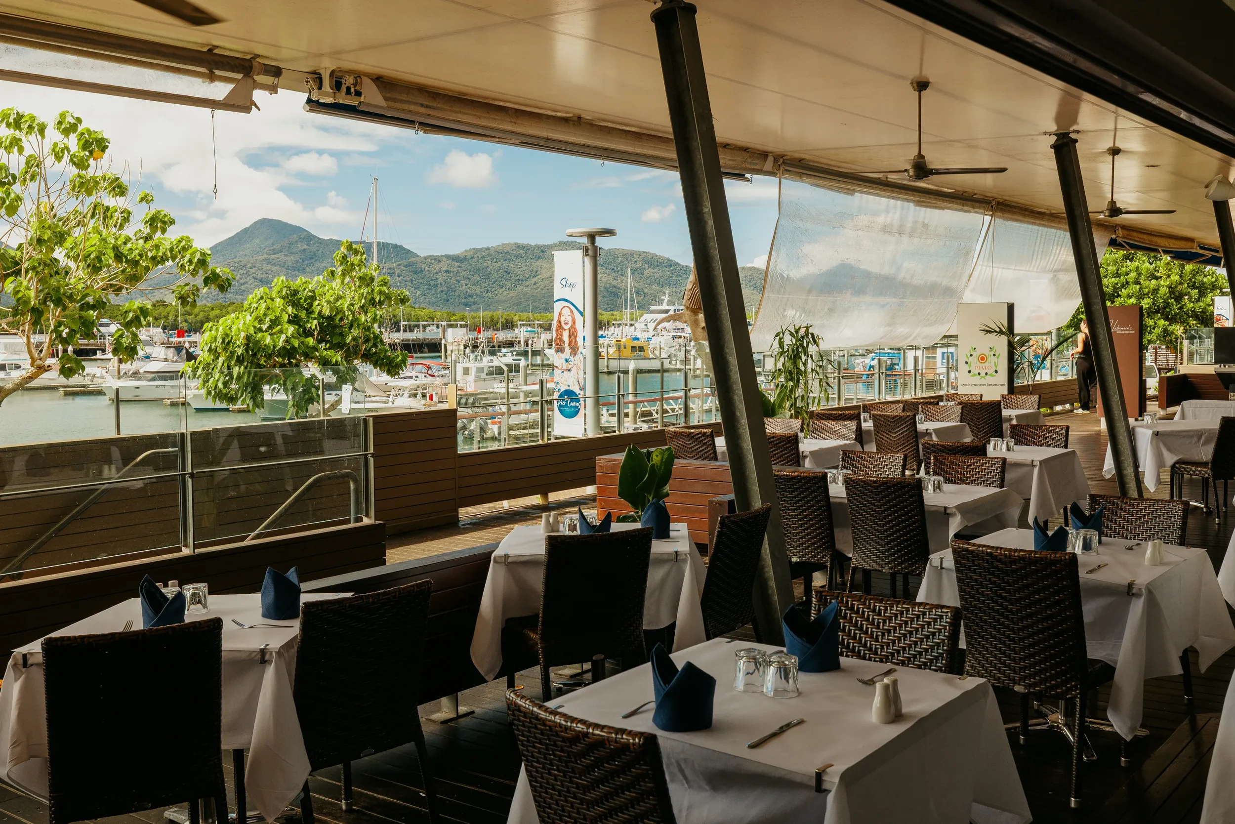 Outdoor restaurant with white tablecloth-covered tables, black wicker chairs, and blue napkins, overlooking a marina with boats and mountains in the background.