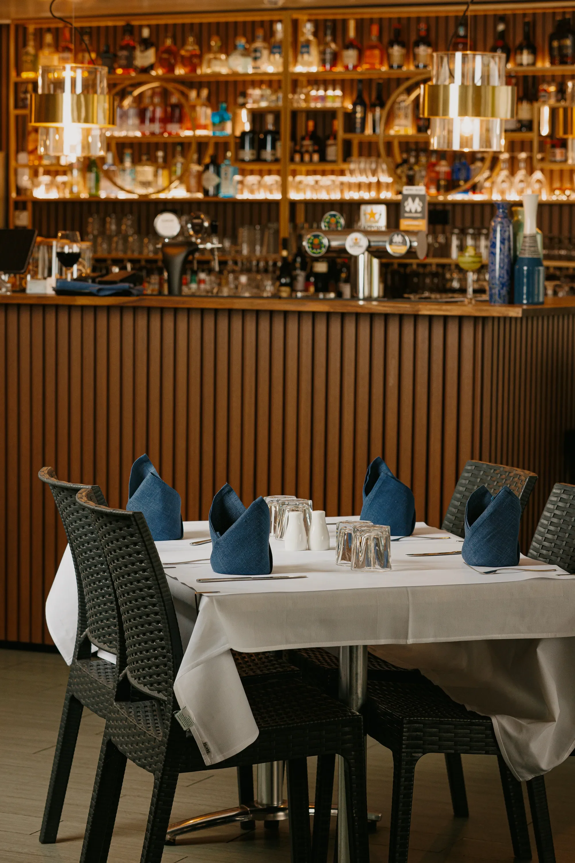 A restaurant dining area with a table set with navy blue napkins, glasses, and cutlery, surrounded by black woven chairs. In the background, there is a bar with shelves of liquor bottles and decorative lighting.
