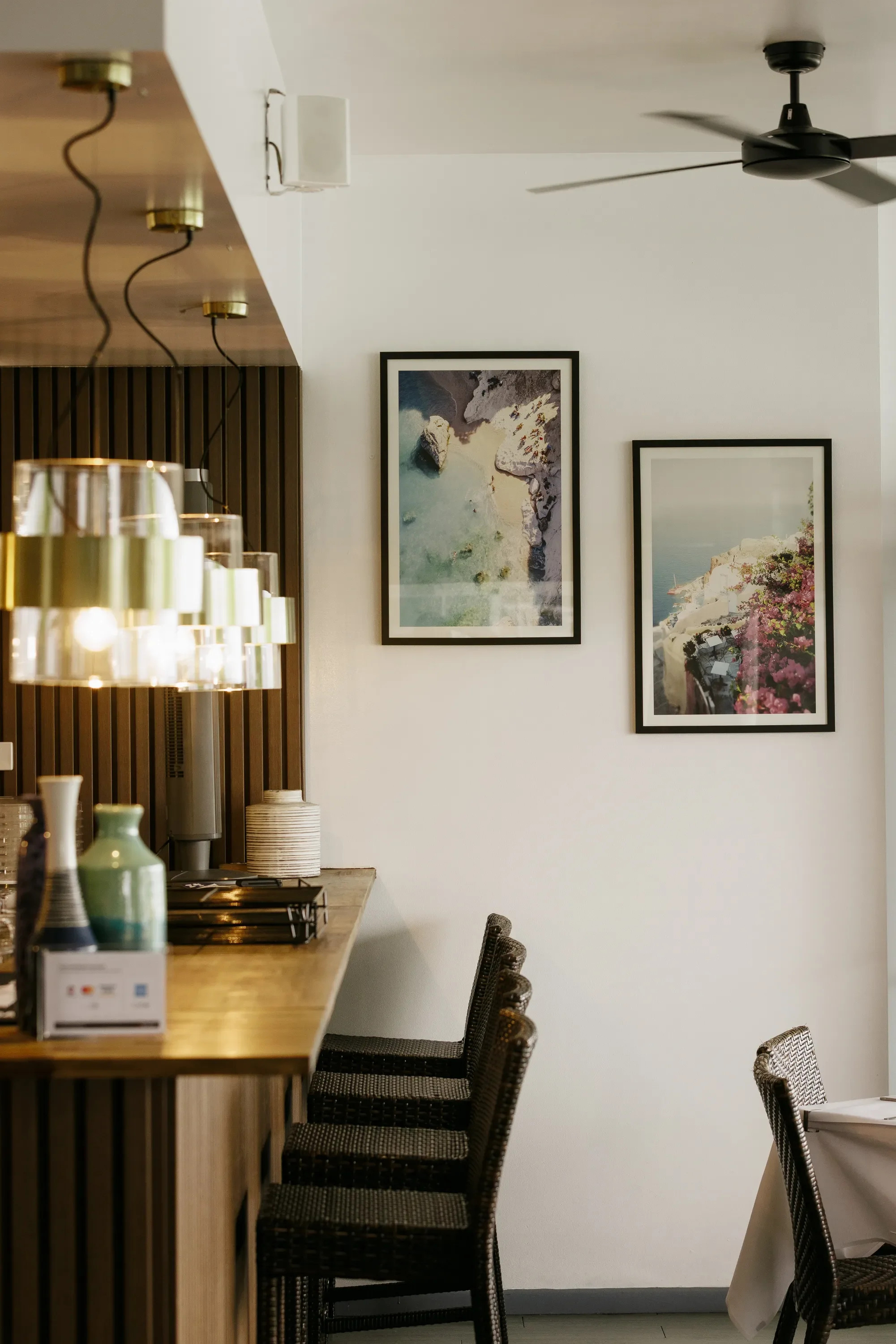 A section of a restaurant or cafe interior with a wooden counter, woven chairs with dark cushions, framed artwork of coastal scenery on a white wall, and a ceiling fan.