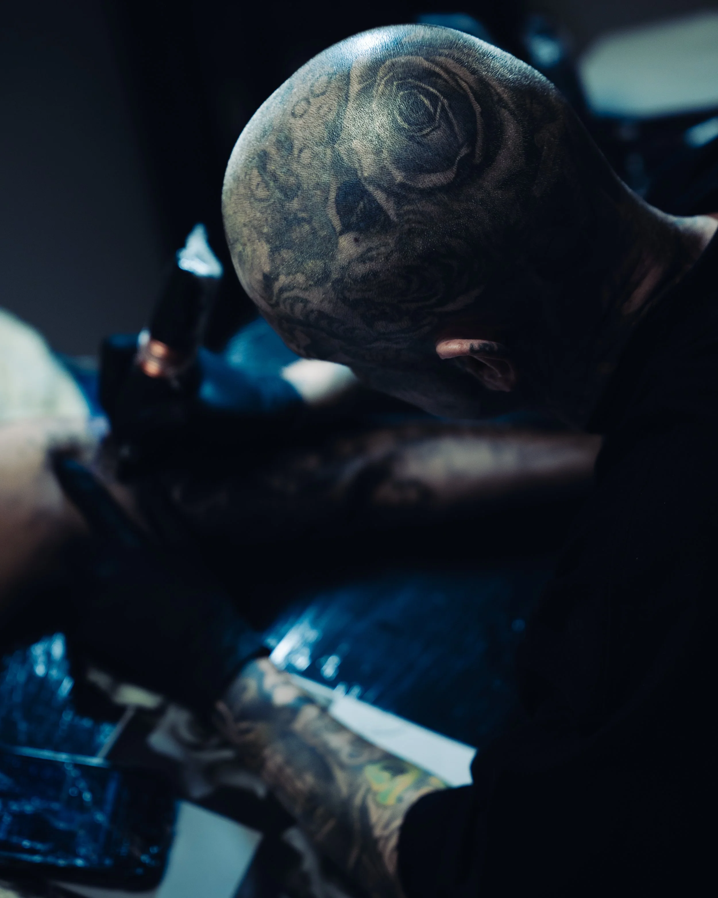 Close-up of a person's head with intricate tattoos, leaning forward in a dark environment with a metallic surface below.