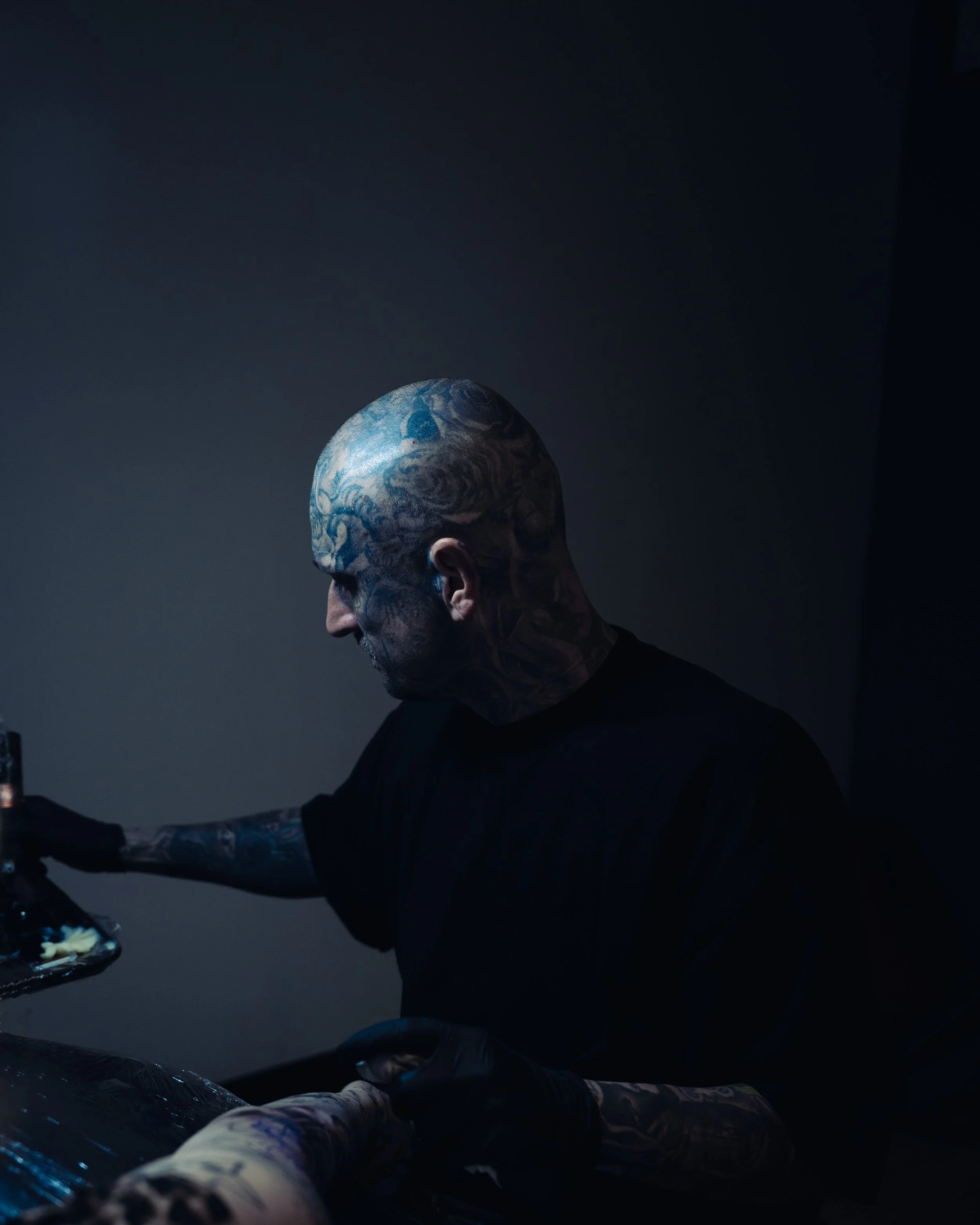 A person with tattoos on their head and arms, wearing a black shirt, is reaching towards a black object with a reflective surface in a dark room with minimal lighting.