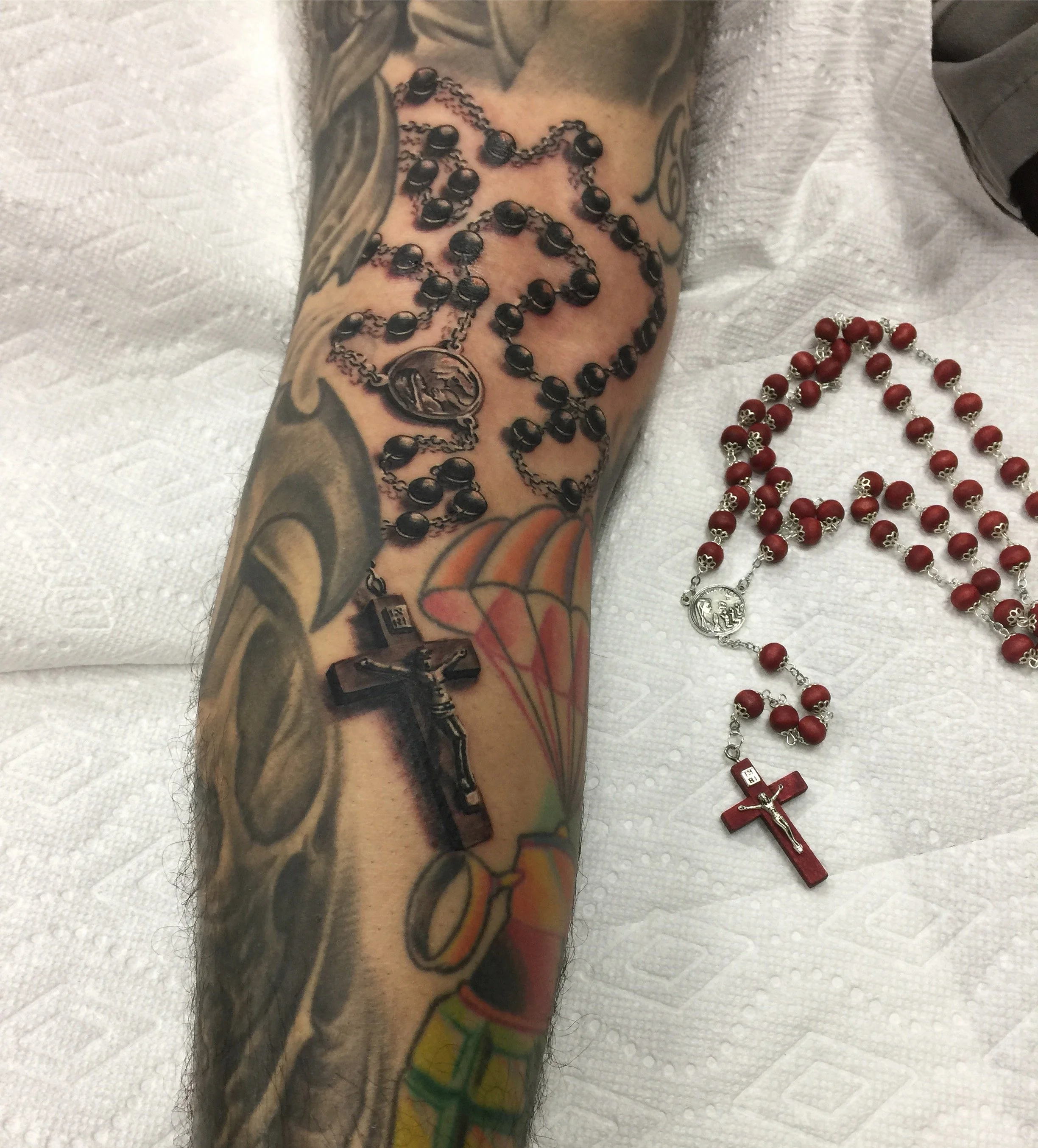 A tattooed arm with various designs including a religious figure and a colorful pattern, resting on a white textured surface. Beside the arm, there is a red rosary with a crucifix and an image of a saint.