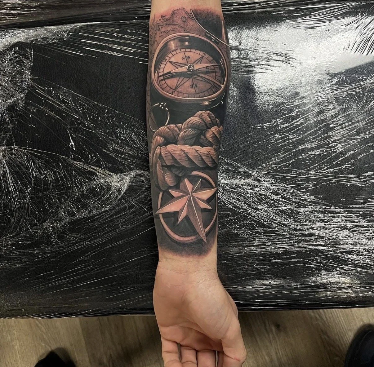 Realistic black and gray tattoo on an arm featuring a compass, a coiled rope, and a star symbol.