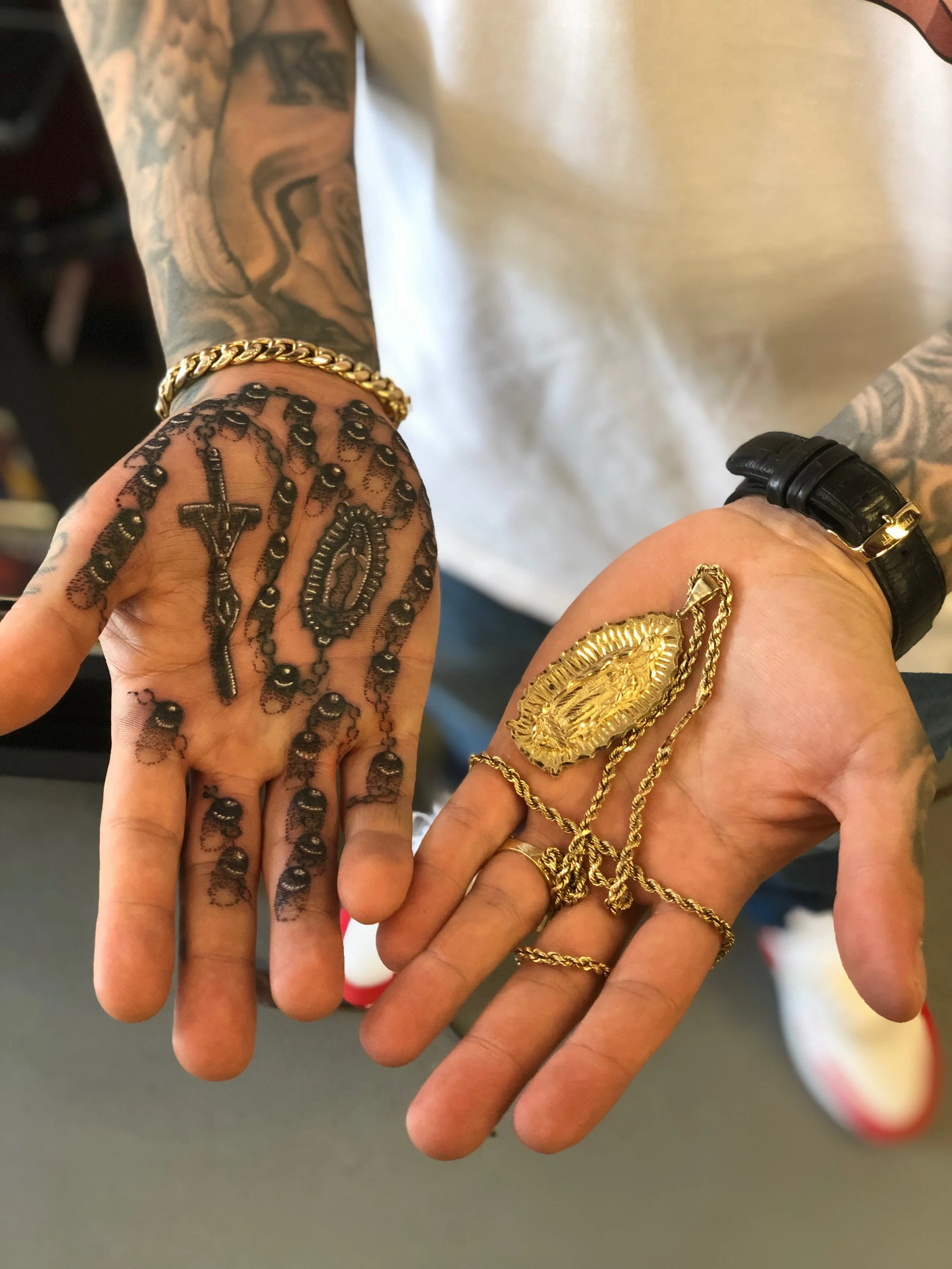 Two hands with tattoos and jewelry, including gold chains and large gold medallions, are depicted in the image.