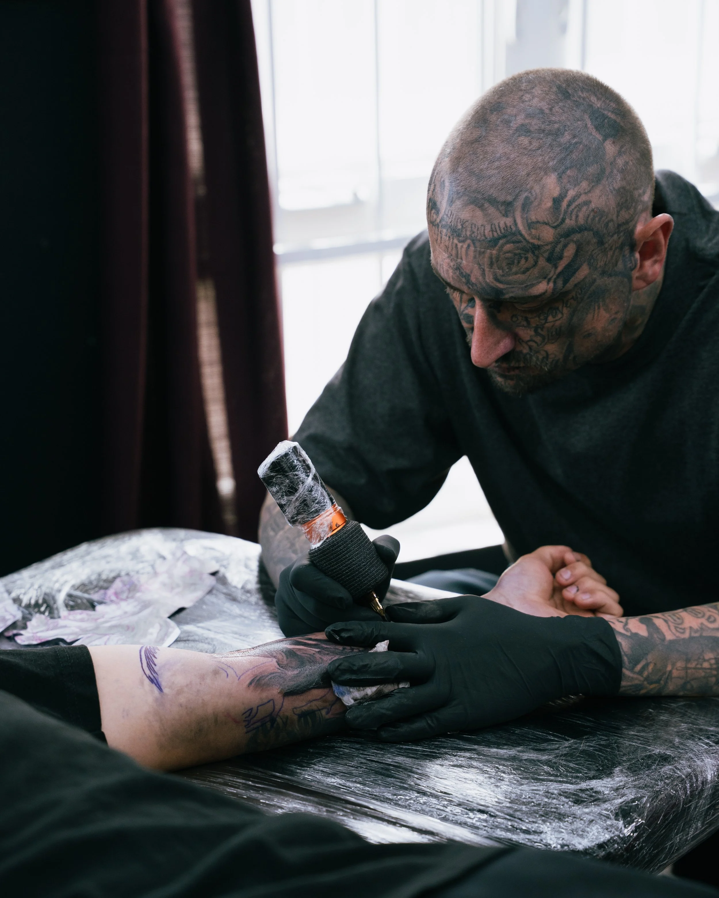 A tattoo artist with tattoos on his head tattoos on his hands and arms is tattooing a person's arm while wearing black gloves, in a tattoo studio with a window in the background.