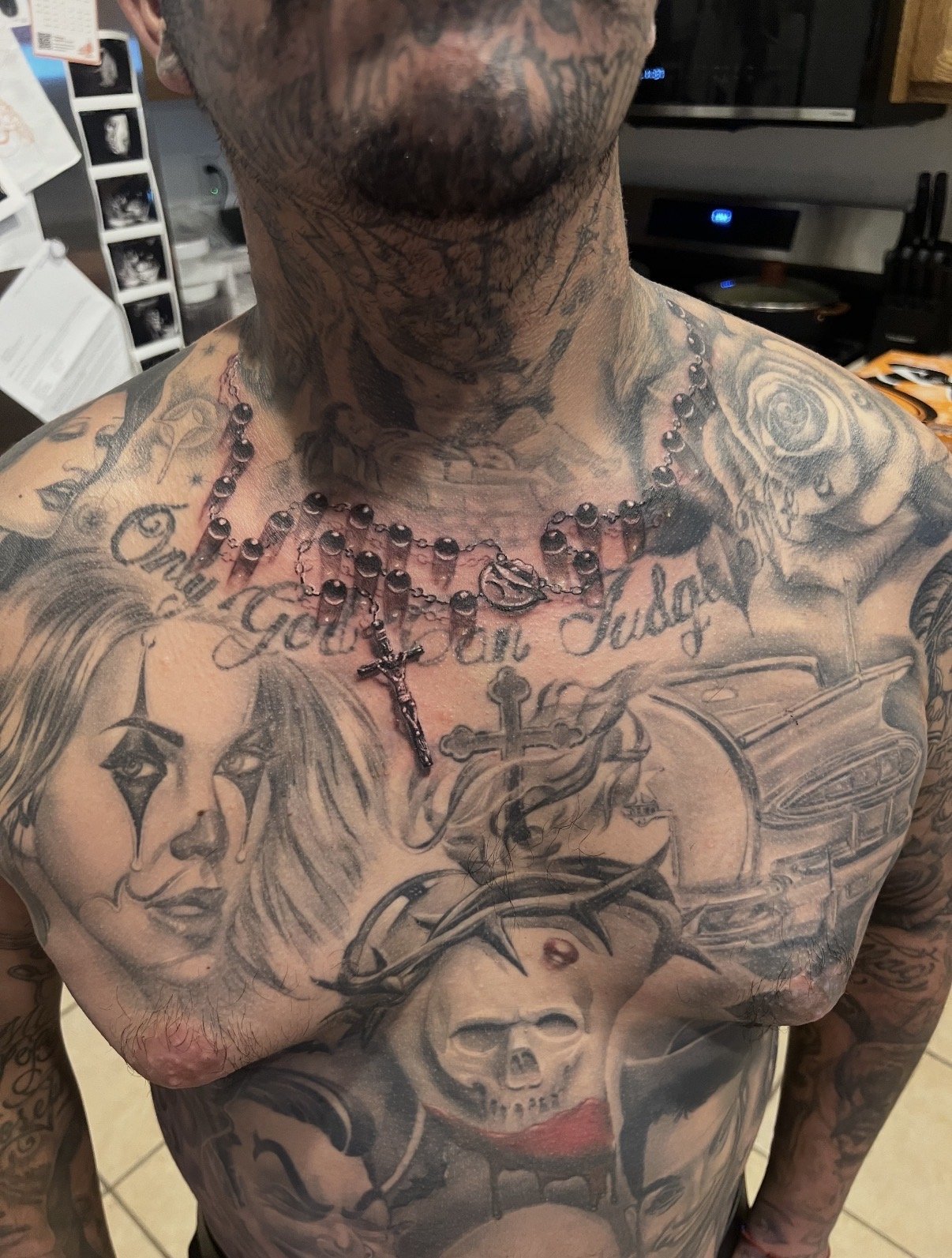 A tattooed man with a large chest and neck tattoos, wearing a rosary necklace with a cross and a heart-shaped pendant. The tattoos include a woman's face, skeleton skull, roses, and various decorative designs.