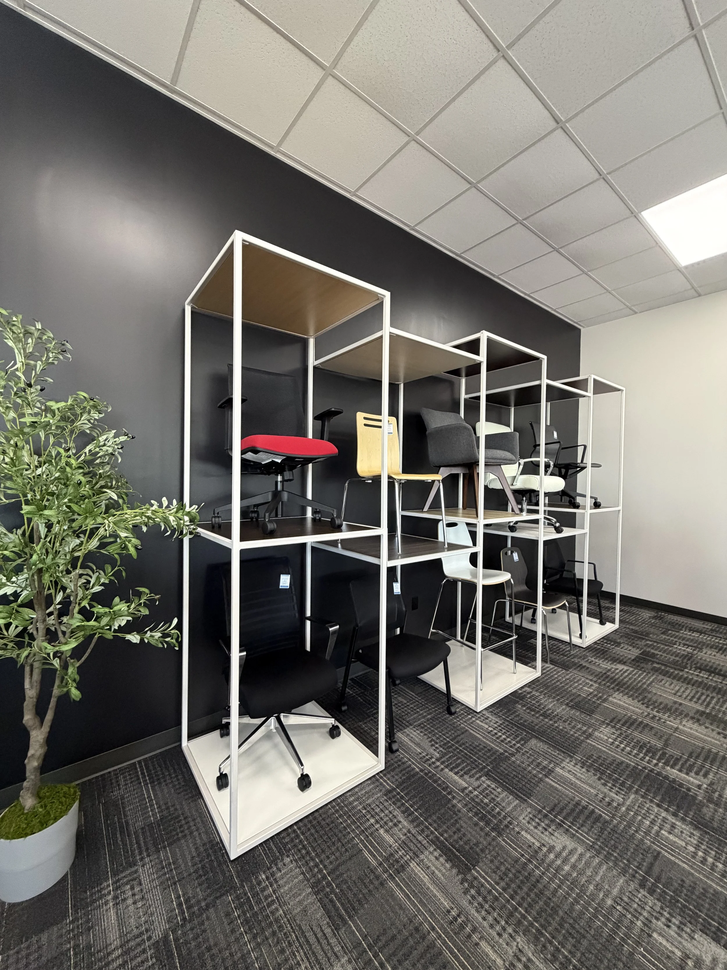 Office space with a black wall, ceiling tiles, and a display of chairs on a white shelving unit, with a potted plant to the left.