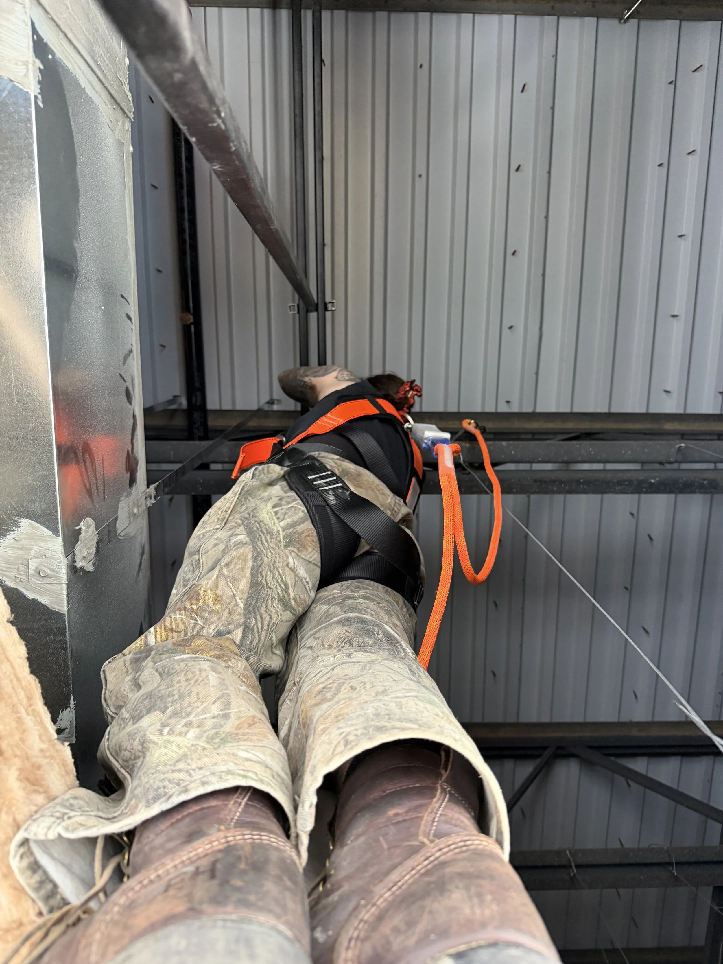 Person lying on a steel beam, wearing camouflage pants, boots, and working at a construction site with a metal roof overhead.