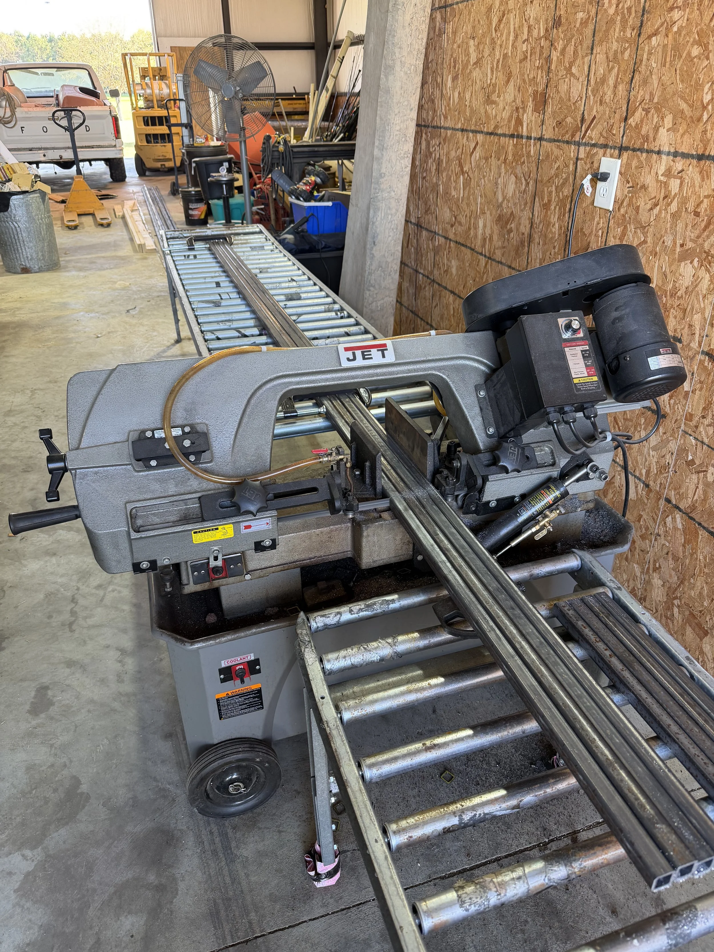 A metal gantry saw with a long cutting track, located inside a workshop with various tools, equipment, and a large electric fan in the background.