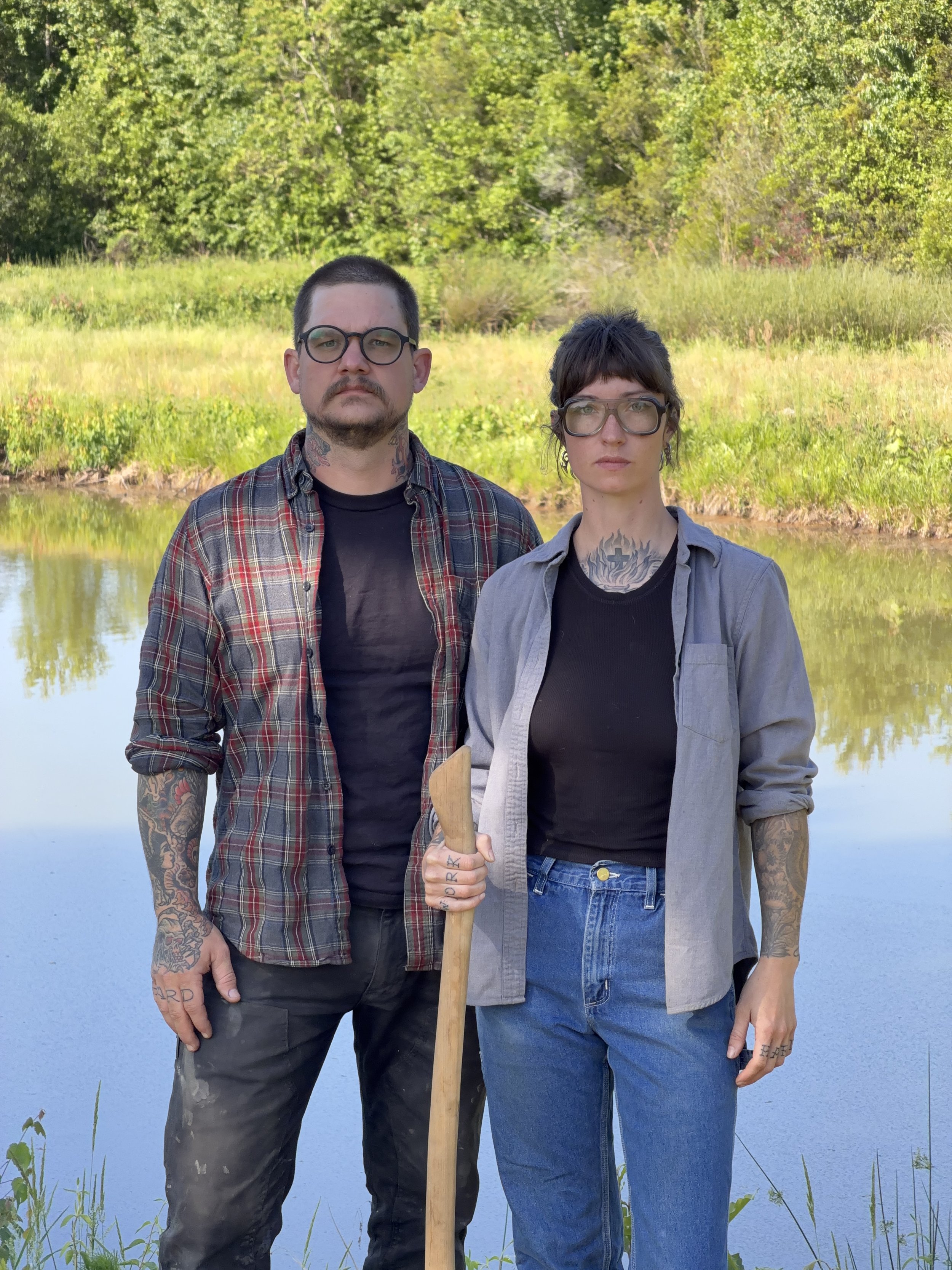 Two people standing outdoors near a body of water with green trees in the background. The person on the left is a man with short dark hair, glasses, tattoos, and wearing a plaid shirt. The person on the right is a woman with short dark hair, glasses, and tattoos, wearing a gray shirt and denim jeans. She's holding a wooden stick. They both have neutral expressions.
