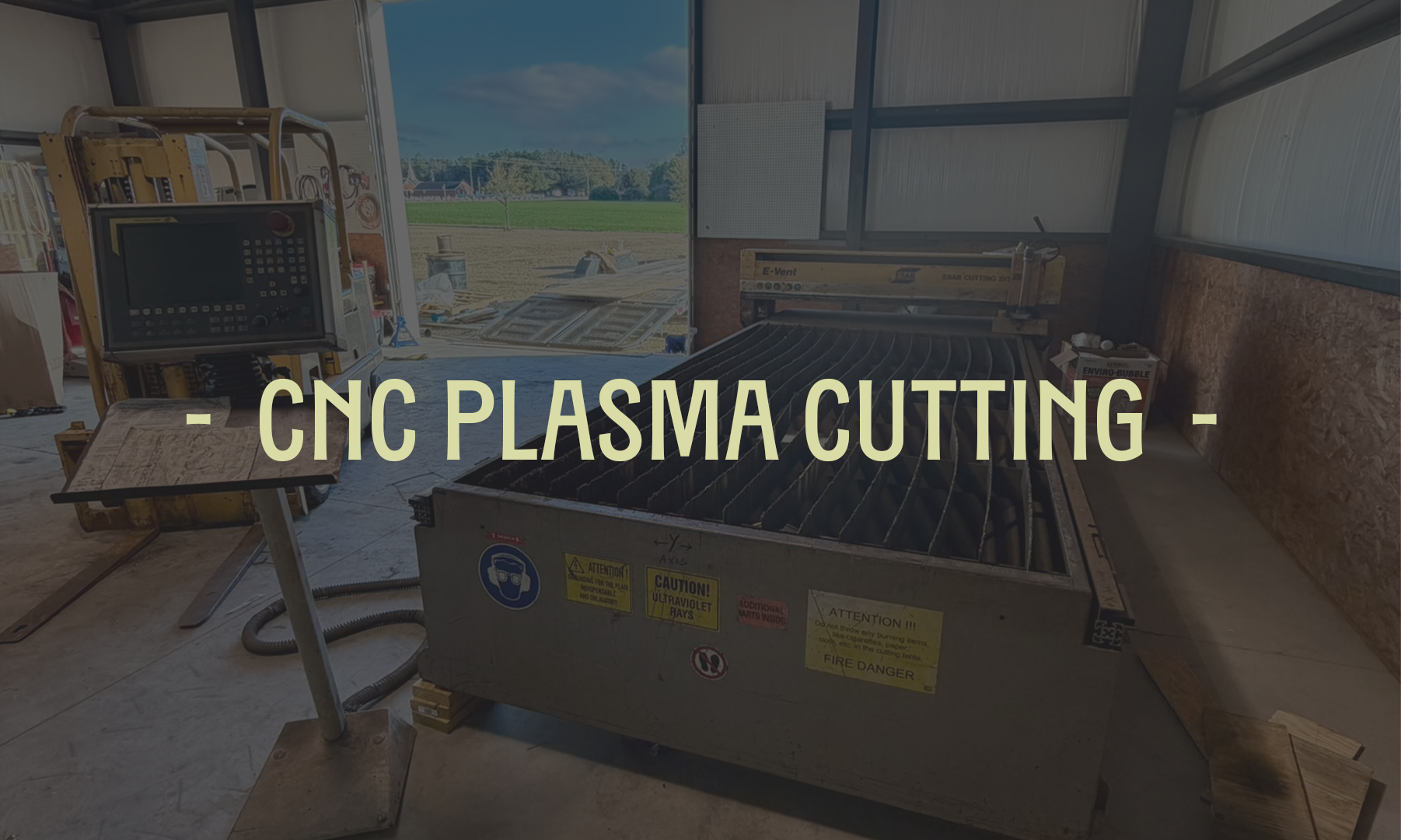 CNC plasma cutting machine inside a workshop with a view of outdoor fields through the open side of the building.
