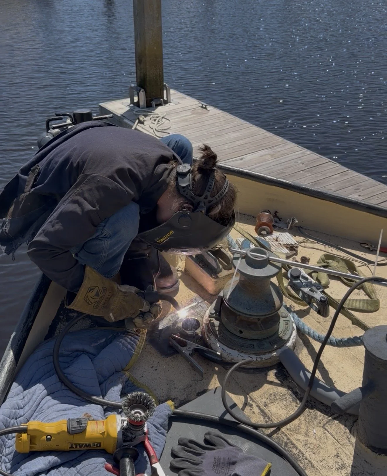 A person welding on a boat near a waterfront dock, with welding equipment and tools visible around.