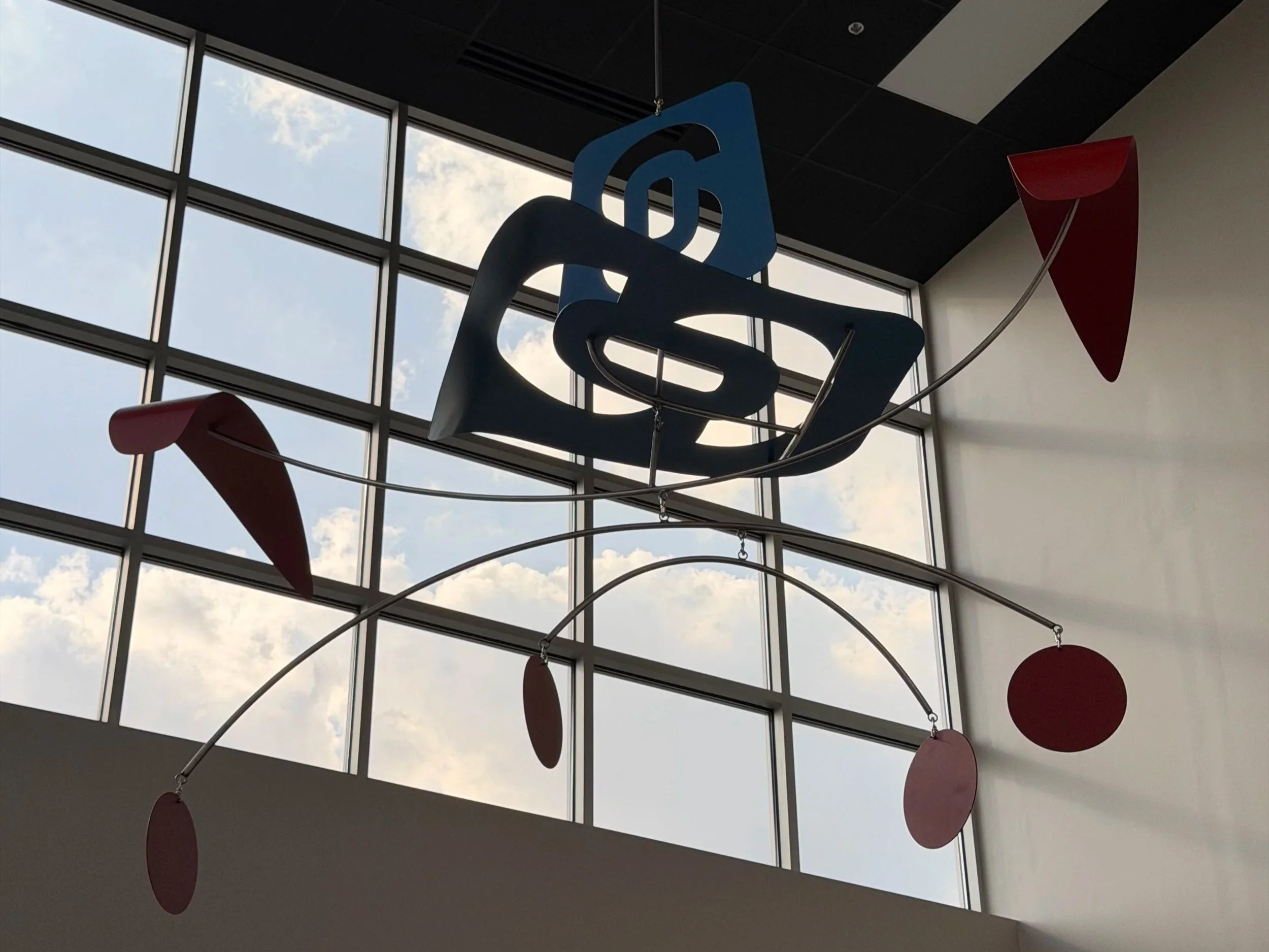 Interior view of a building with large glass windows showing a blue sky with clouds, decorative hanging elements including large red circles and red cone-shaped objects, and a blue sign with black text, possibly part of a logo or artwork.